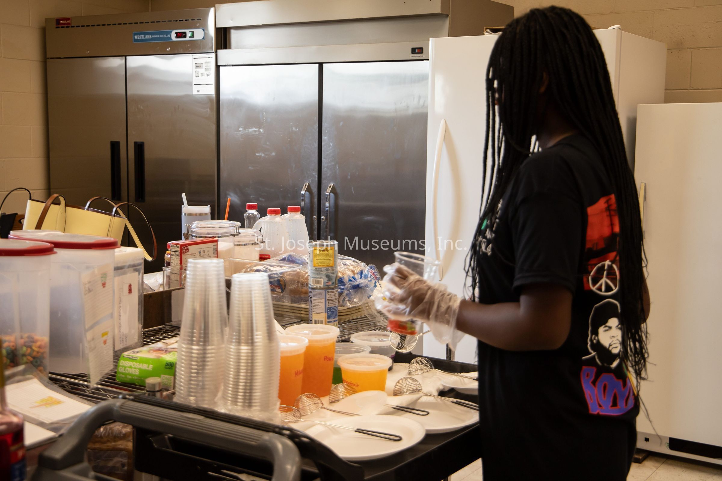 In the image, a person with long braided hair is standing in a kitchen area, engaged in preparing or organizing food items on a counter. The individual is wearing a black t-shirt with colorful graphics and is using disposable gloves, indicating an emphasis on hygiene. The countertop is crowded with various items, including clear plastic cups, containers of juice, canned goods, and bread. These items are neatly arranged, suggesting a setup for serving food or drinks. Behind the person, there is a large stainless steel refrigerator, along with another, smaller white one, hinting at a well-equipped kitchen environment. The scene conveys a sense of busy activity, perhaps in preparation for a group event or meal service.