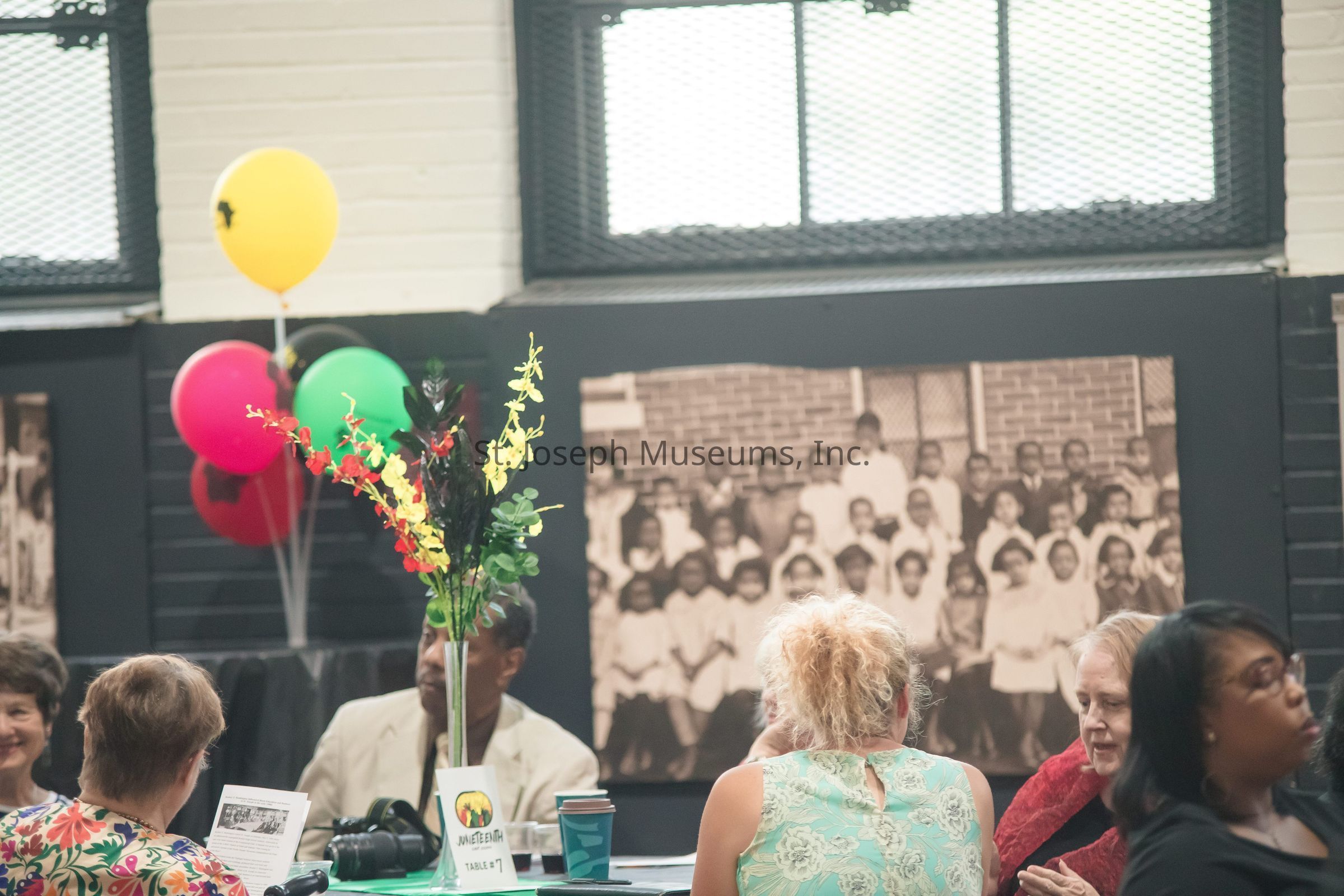 In this lively image, a group of people is gathered around a table covered with a green tablecloth, engaging in conversation at what appears to be a social event or gathering. The table is adorned with a vibrant centerpiece featuring tall, colorful flowers in red, yellow, and green hues, which complements the festive atmosphere. In the background, a large black-and-white photograph of a group of people is displayed on the wall, adding a sense of history or nostalgia to the setting. A cluster of balloons in bright red, green, and yellow, tied near the table, suggests a celebratory occasion. The attendees, dressed in casual to semi-formal attire, seem relaxed and engaged, indicating a welcoming and communal environment. The room's decor, including the arrangement of chairs and the presence of a camera on the table, hints that this might be a special reunion, cultural gathering, or commemorative event. Large windows with grid mesh allow natural light to filter into the space, further illuminating the scene.