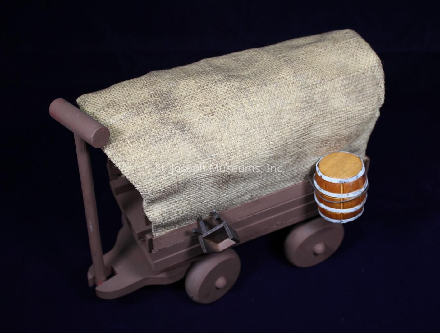 The image displays a model of a traditional covered wagon, often associated with historical American pioneer life. The wagon is crafted with a rustic brown wood appearance and features a curved, beige fabric cover that evokes the style of the covered wagons used during westward expansion. It rests on four wooden wheels that are firmly attached, giving it a robust appearance. A small wooden barrel, detailed with metal bands, is fastened to one side of the wagon, suggesting storage or transport of goods. The setting is simple, allowing the focus to remain on the wagon's details.