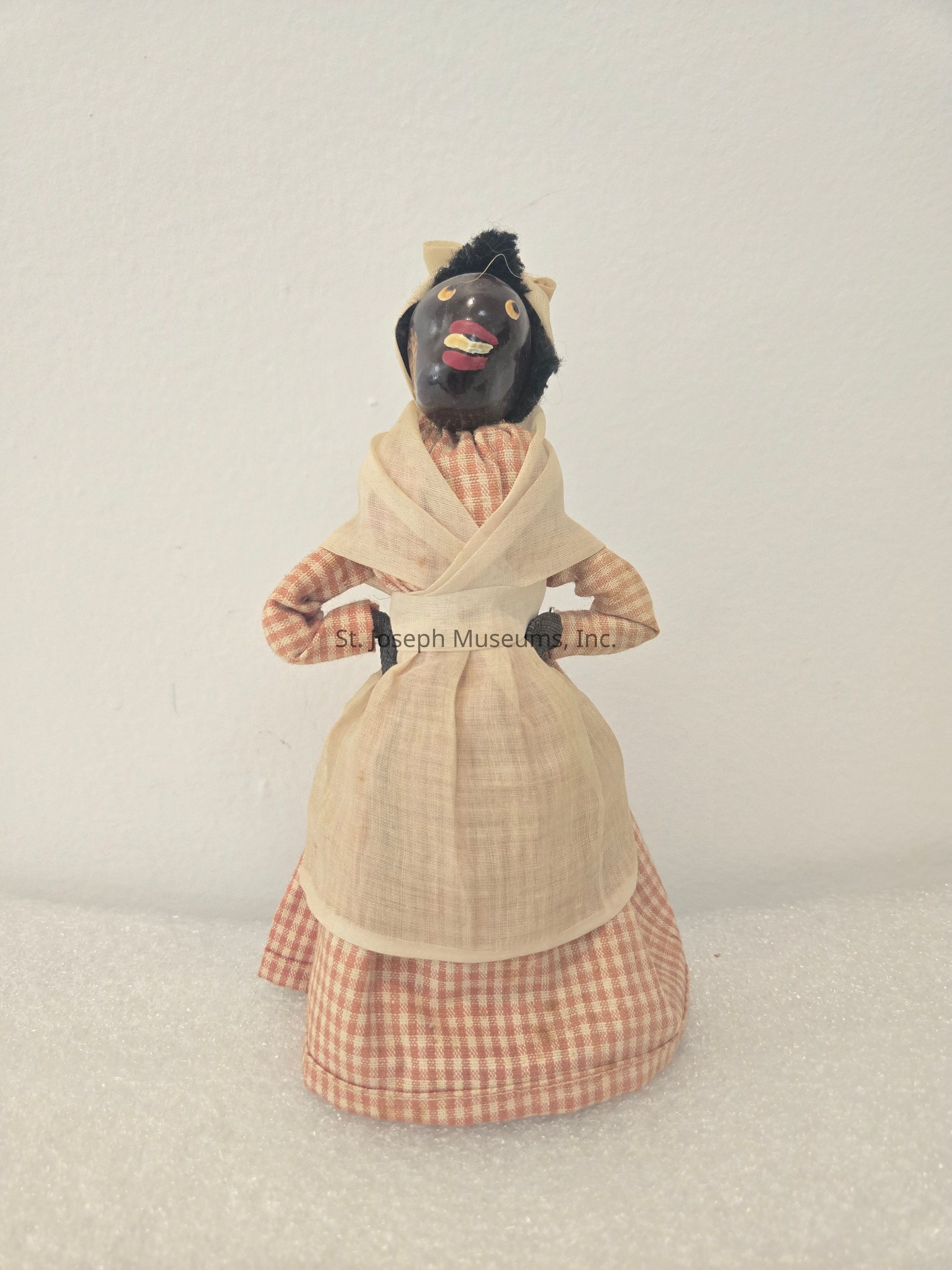 The image features a vintage doll dressed in a traditional outfit that consists of a checkered dress with a long skirt and a cream-colored apron and shawl wrapped around its shoulders. The doll has a quirky, expressive face with wide eyes, a broad smile, and a headscarf tied around its head. Its hands rest on its hips, giving the impression of a confident stance. This style of doll reflects an antique or historical design, showcasing the craftsmanship of a past era. The background is plain, allowing the focus to remain entirely on the doll, highlighting both its attire and its detailed features.