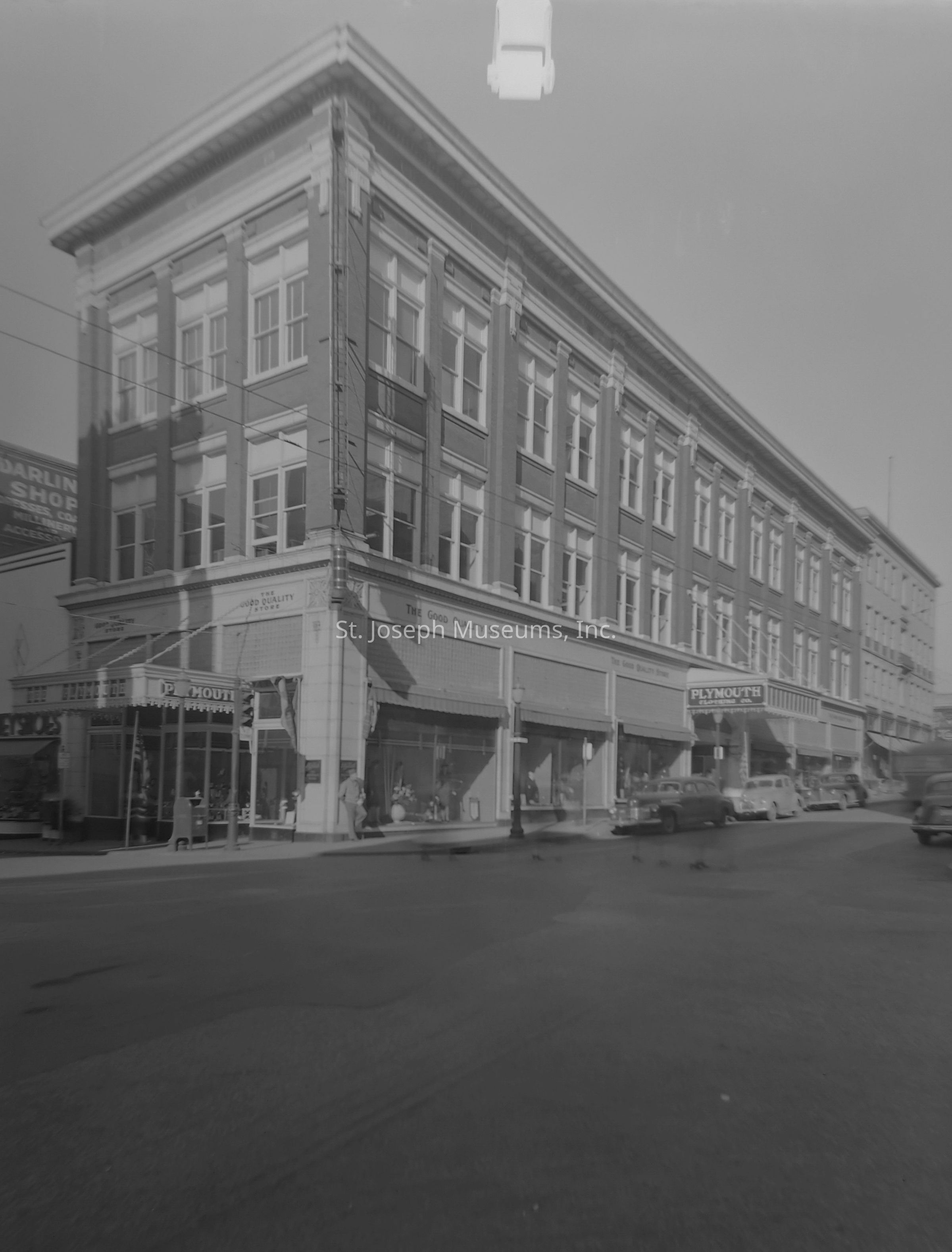 Black-and-white image showscasing the exterior of the Plymouth Clothing Co. located at the northwest corner of 6th and Felix in St. Joseph, Missouri, featuring a classic three-story brick building with large windows and intricate architectural details. Several vintage cars are parked alongside the street, and people can be seen walking near the storefronts, which display clothing items. The signage prominently showcases the name of the clothing company, emphasizing its identity as "The Good Quality Store."