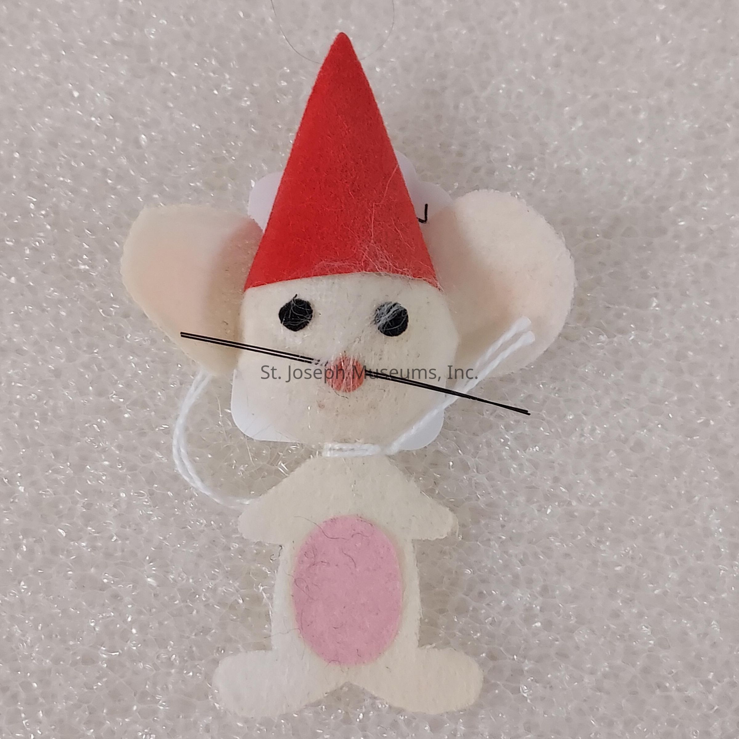 The image showcases a cute, handcrafted felt mouse ornament with large, round ears and an expressive face composed of small black eyes, a pink nose, and long, thin whiskers. The mouse is dressed in a vibrant red pointed hat atop its head, adding a whimsical and festive flair to its appearance. Its body is primarily an off-white color, with a soft pink oval on its belly, further enhancing its endearing look. The mouse is set against a textured background of white bubble wrap, which provides a simple yet effective contrast. The delicate craftsmanship and charming design suggest that this ornament would make a delightful addition to holiday decorations or a festive display. The lightweight material and hanging loop indicate its easy placement on a Christmas tree or in other decorative settings. This adorable little mouse embodies a sense of playfulness and charm that can bring a touch of joy and warmth to any festive occasion, making it a perfect decoration for anyone who appreciates handcrafted, whimsical designs.
