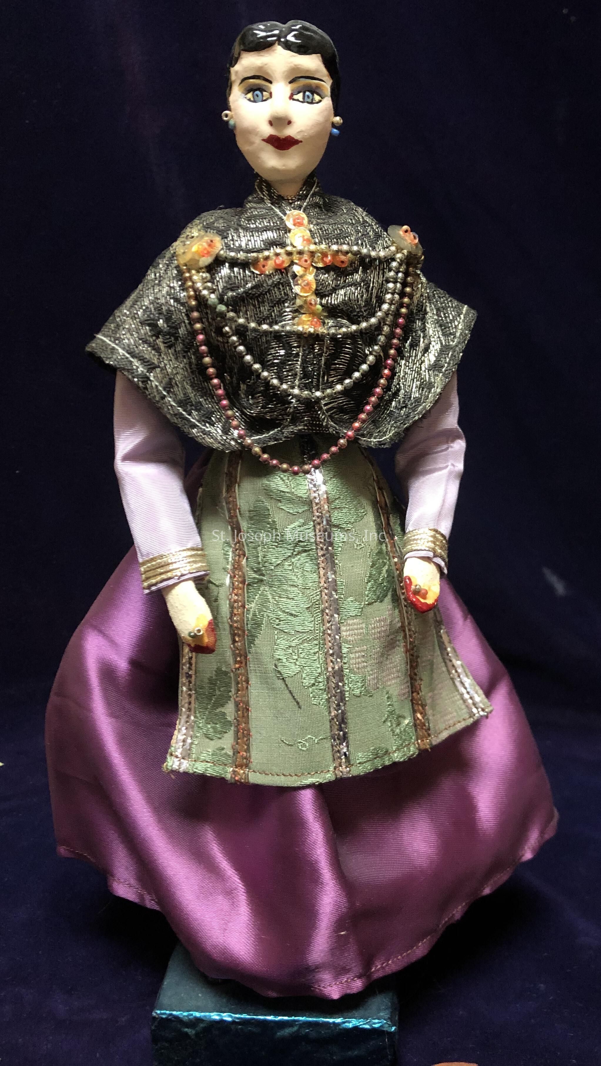 The image features a female doll with an intricately painted face, adorned with vivid lipstick and eye shadow, and crowned with dark, glossy hair. The doll is dressed in an elaborate outfit comprising a lush, purple satin skirt and a light green, patterned overlay, enhancing her stately appearance. The ensemble is complemented by a metallic, embroidered shawl draped elegantly over her shoulders, intricately detailed with vibrant beads and gems that cascade down in strands. Her hands, delicately sculpted and dressed in coordinating cuffs, rest calmly at her sides, while the entire figure stands poised on a small, dark platform against a deep blue backdrop. The attention to detail in the craftsmanship suggests cultural or traditional significance, offering a glimpse into an artistic interpretation of historical fashion.