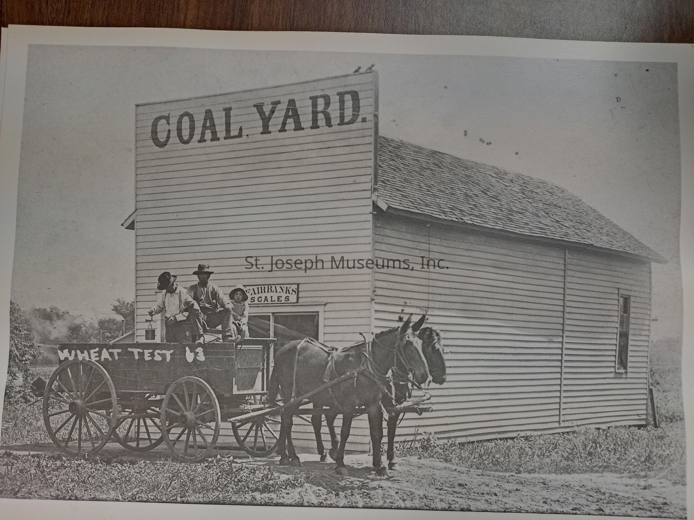 Black-and-white image depicting an old-fashioned coal yard with a large wooden building labeled "COAL YARD" and a sign for "FAIRBANKS SCALES" on its side. In front of the building, a horse-drawn wagon with the words "WHEAT TEST 63" painted on its side stands on a dirt path. Three men sit on the wagon, two in hats and casual work attire, under the open sky.