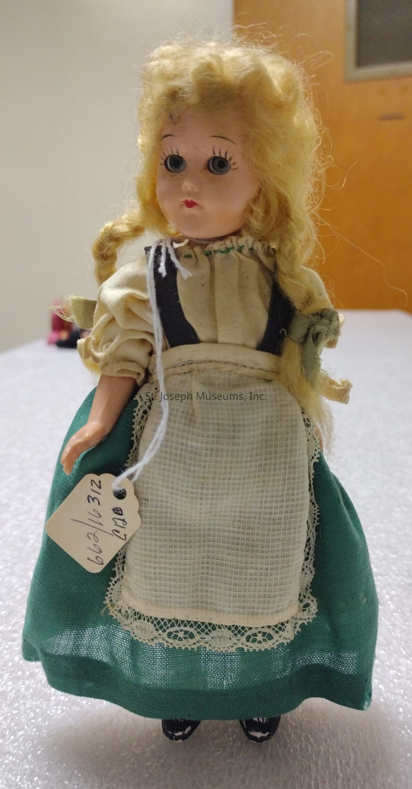 The image shows a vintage doll with curly blonde hair and large blue eyes, dressed in traditional attire featuring a cream-colored blouse with puffed sleeves and a green skirt adorned with a white lace-trimmed apron. The doll has a slightly open mouth, giving it a quaint expression. It is standing upright on a textured white surface, possibly a table, and there is a small tag attached to its arm with handwriting that appears to have a number or code. In the background, a beige wall and a wooden door can be seen, indicating it might be in an indoor setting, such as a collection or museum display. The doll's shoes are black with decorative detailing, completing its charming appearance.