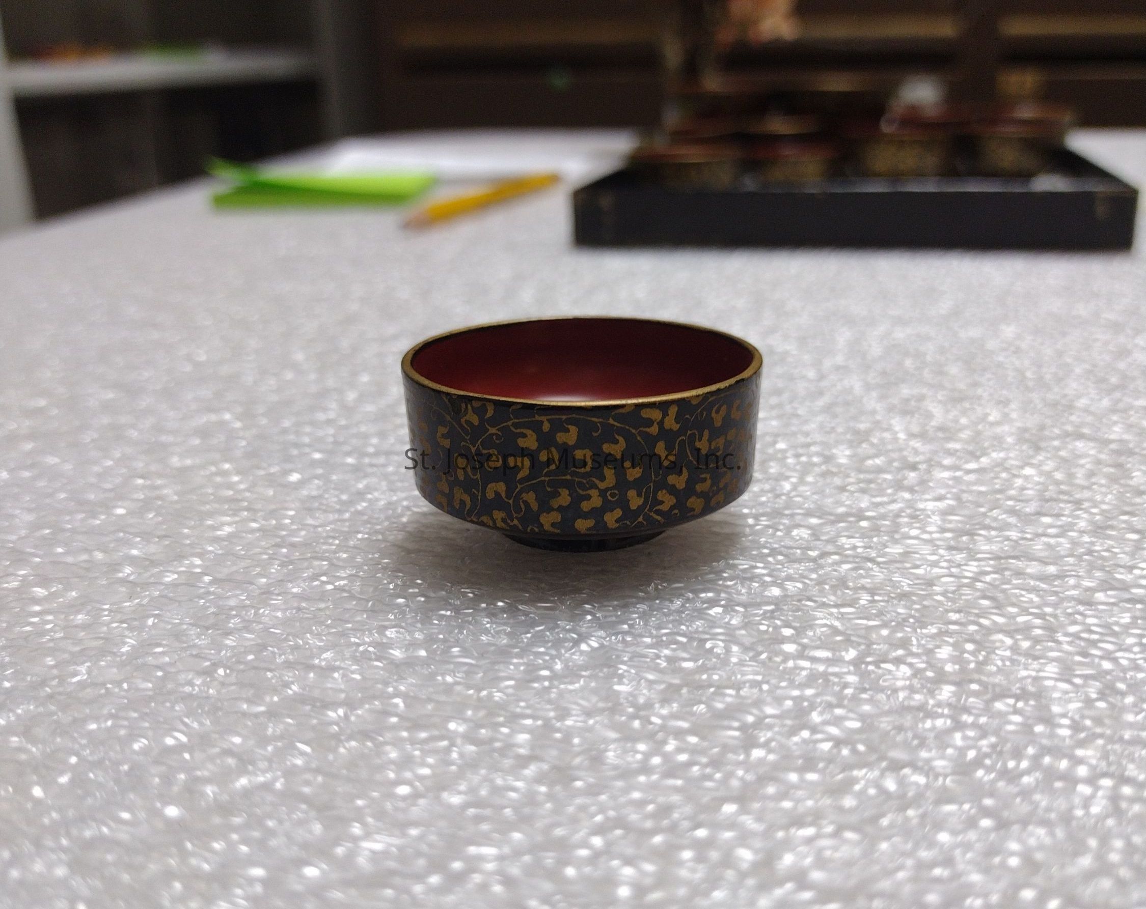 The image depicts a small, ornate cup with a rich, deep red interior and a black exterior adorned with an intricate pattern of gold designs, creating an elegant and sophisticated appearance. This cup is placed on a surface covered with bubble wrap, adding a textural contrast between the delicate cup and the protective layer underneath. In the background, slightly blurred, there are other similar cups on a tray, suggesting a set, along with a yellow pencil and some green paper. The setting appears to be a table or workspace, hinting at a context of organization or preparation, possibly for a cultural or tea-related event. The overall composition of the image implies an attention to detail and an appreciation for artistry and craftsmanship, as reflected in the cup's design and the careful arrangement of surrounding items, which contributes to a visually interesting and harmonious scene.