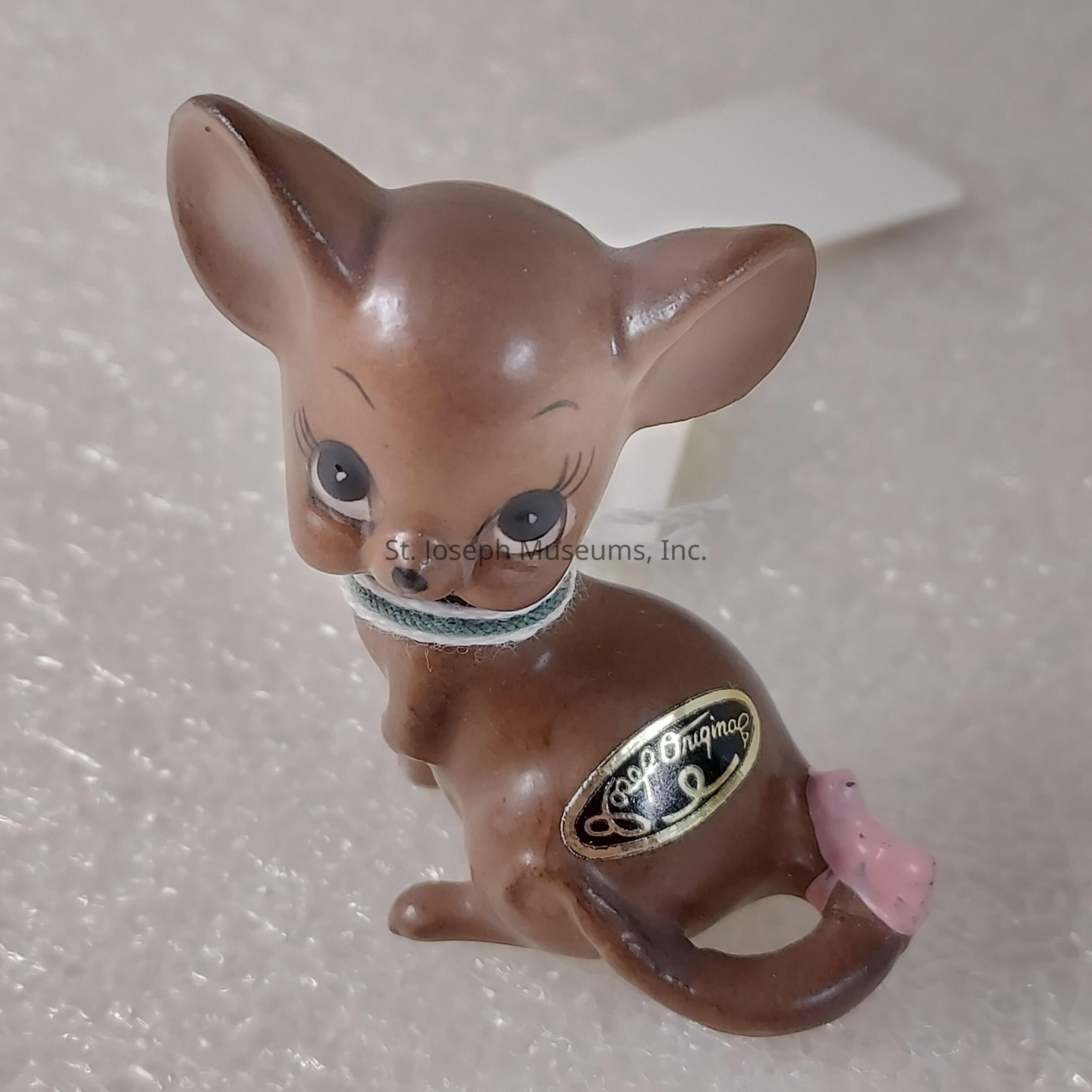 The image depicts a small, brown ceramic figurine of a baby deer with large, expressive black eyes and oversized ears, presenting an adorable and innocent appearance. A subtle, light-colored string encircles its neck, possibly acting as a marker or decorative element. The figurine's smooth, glossy surface reflects light, highlighting its fine craftsmanship. Attached to the back, there is a golden sticker that reads "Josef Originals," suggesting the piece belongs to a well-known line of collectible items. A small, pink bow adds a charming touch to the figure's tail, contributing to its endearing and whimsical character. The figurine is positioned against a textured, light background, which enhances its features and makes the deer stand out prominently. Its pose, with a gentle, sitting posture and attentive look over its shoulder, evokes a sense of curiosity and playfulness, capturing the essence of a young fawn in a moment of pause, ready to explore its surroundings.