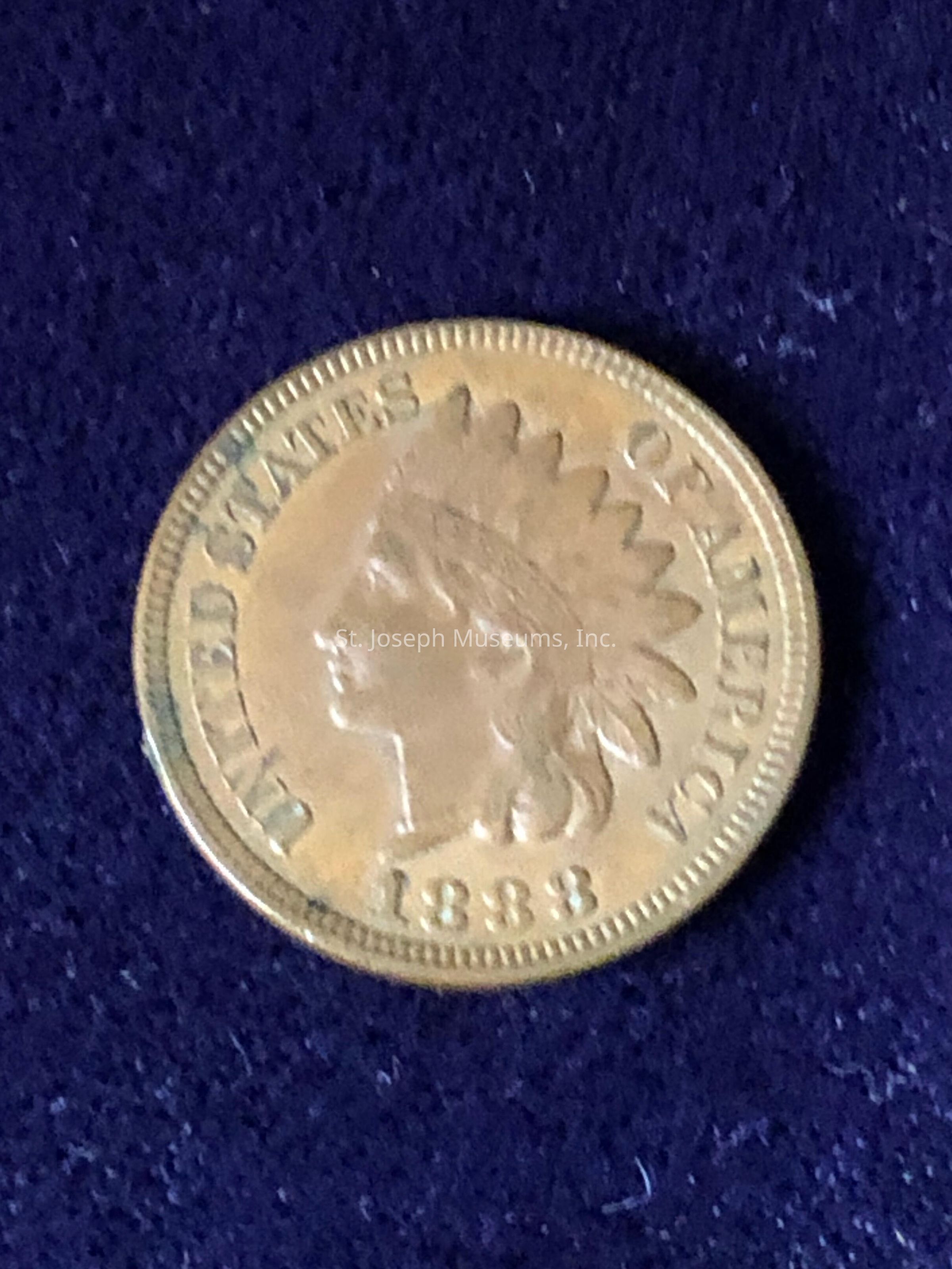 The image shows a vintage coin with a design featuring the profile of a person wearing a feathered headdress, often associated with the United States' "Indian Head" penny, minted in the year 1883. The coin is inscribed with the words "United States of America" surrounding the profile, which is prominently positioned at the center. The coin's surface appears to have a reddish-golden hue, reflecting its metal composition and the age that adds to its historic charm. Its circumferential rim is delicately detailed, enhancing the aesthetic appeal and craftsmanship of the period. The image background is a textured, deep blue fabric that contrasts with the coin, drawing attention to the intricacies and aged elegance of this numismatic piece.