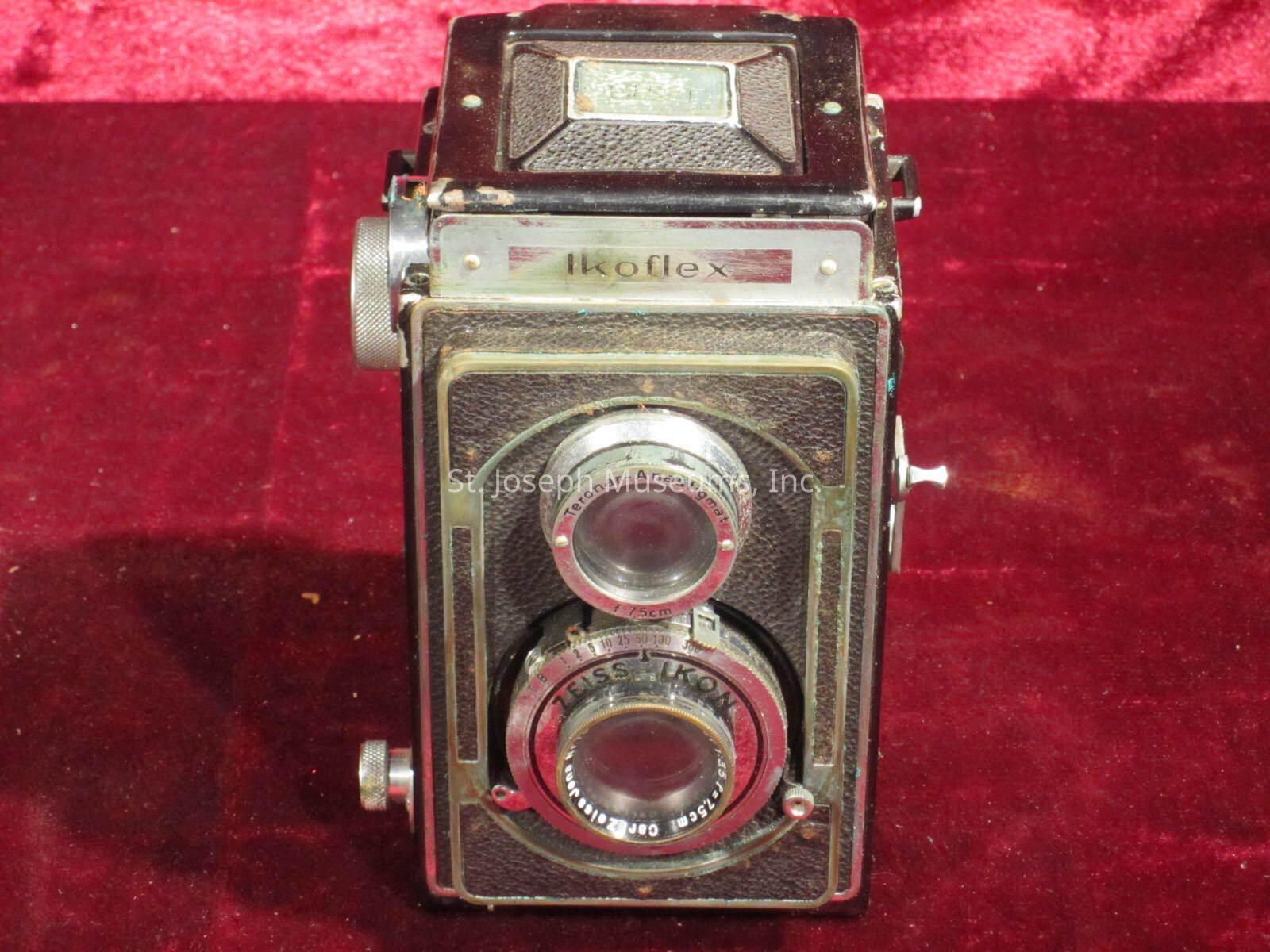 The image shows a vintage Ikoflex twin-lens reflex camera resting on a rich red velvet background. The camera has a classic design with two prominent lenses, surrounded by metal and leather accents, which highlight its retro look. The top of the camera displays the name "Ikoflex," suggesting its brand and model. The textured leather finish, coupled with metal detailing, gives the camera an elegant and durable appearance. The overall design reflects the era of its manufacture, likely mid-20th century, a time when such cameras were popular for their durability and photographic quality. The red velvet adds a luxurious and nostalgic feel, emphasizing the camera's status as a collectible or cherished antique.