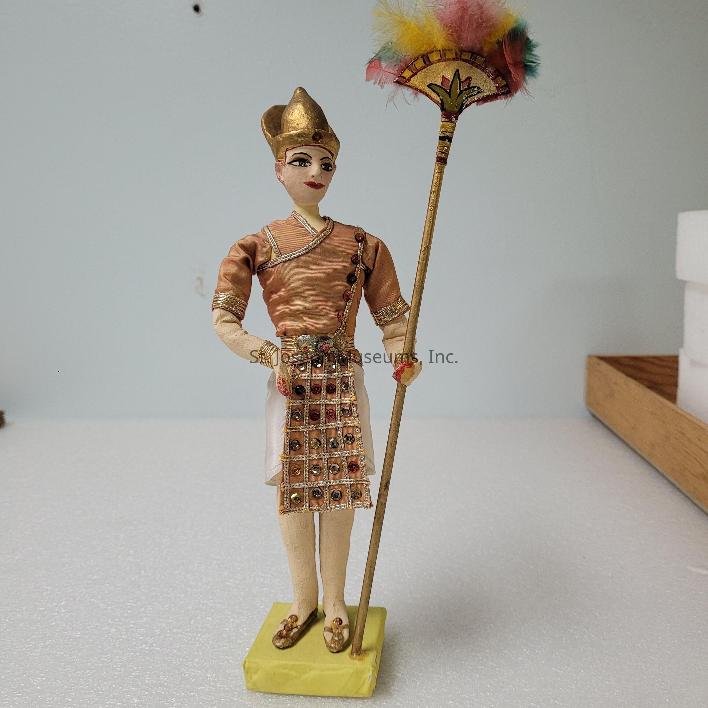 The image features an intricately designed figurine standing on a yellow base. The figure is dressed in traditional attire, including a detailed tunic adorned with decorative patterns and beading, giving it an ornate appearance. It holds a slender staff topped with colorful feathers, adding vibrant hues to the composition. The figure wears a golden metallic headdress, enhancing its regal look. The figurine's expression is poised, with subtle facial details painted meticulously. Its sandals and accessories complement its overall aesthetic, reflecting a cultural or historical theme. The background is minimalist, with a pale wall and some wooden and foam elements, which places emphasis on the figurine itself. The craftsmanship suggests careful attention to detail, from the clothing's texture to the facial features, highlighting both artistry and cultural significance, making the piece appear both elegant and timeless.