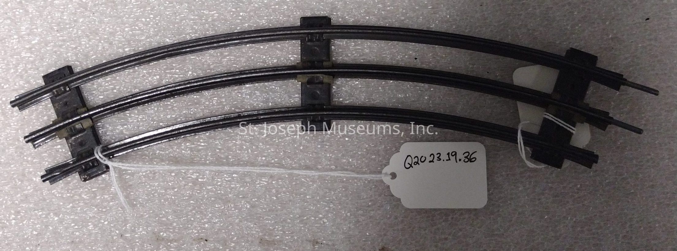 The image shows a pair of curved, metal train track pieces, each comprising two rails held together by several cross ties, creating a gentle arc. The track segments are resting on a textured, light-colored surface that resembles foam or a soft material. Attached to the tracks is a white string, which holds a label with the code “Q20 23.19.36” written in black ink, indicating some form of identification or cataloging system. The scene is well-lit, highlighting the slight shine and smooth texture of the metallic rails, suggesting they are part of a model train set or similar assembly designed for use on a miniature scale, serving as components for constructing intricate layouts or scenes for model trains, and likely collected or cataloged with care using the tag for reference or inventory purposes.