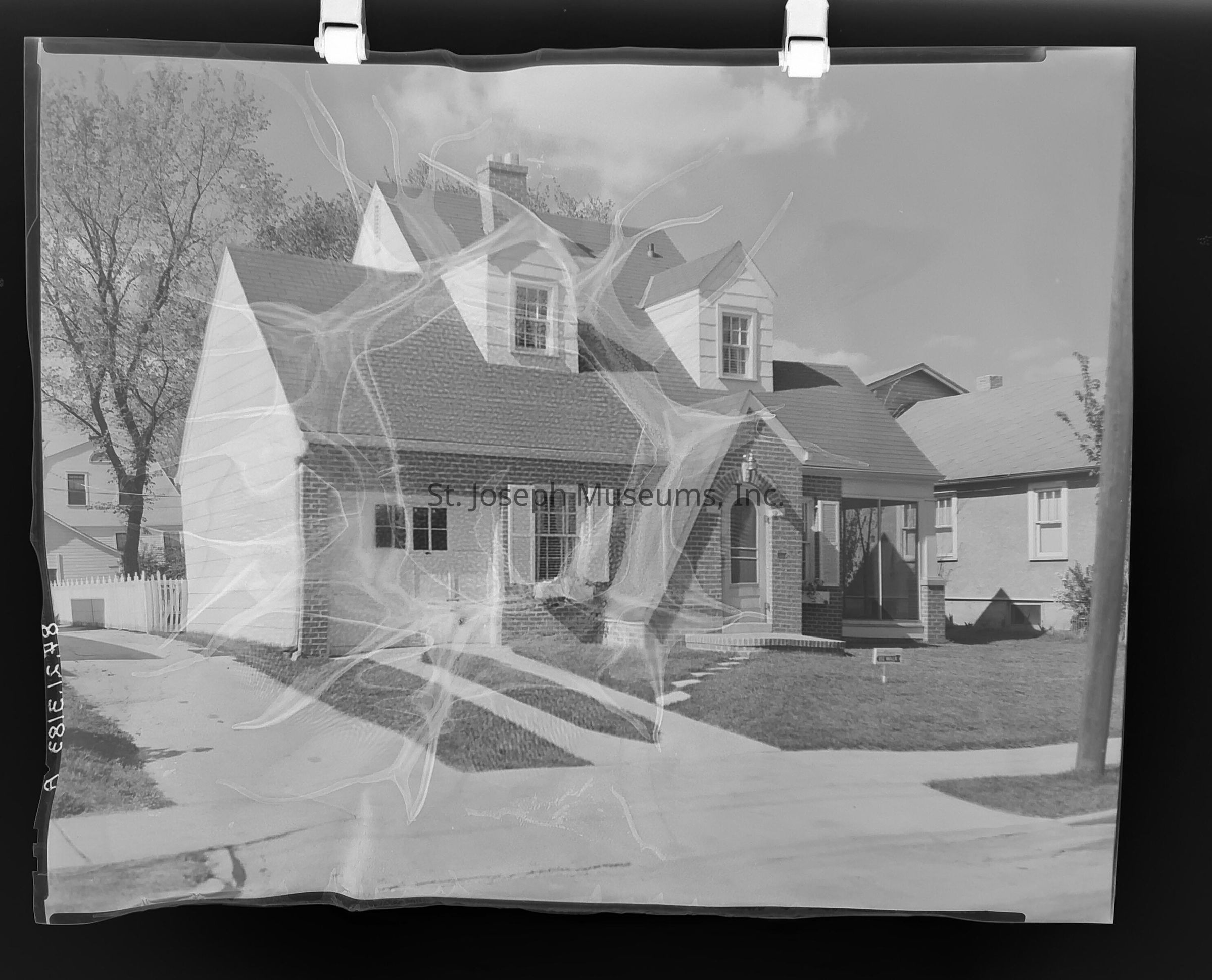 Black-and-white warped photograph showcasing a charming brick house on 2821 Jules Street in St. Joseph, Missouri, featuring a gabled roof, an inviting arched doorway, and shuttered windows. It has a neatly maintained lawn, a driveway leading to an attached garage, and a cozy front porch surrounded by lush trees.