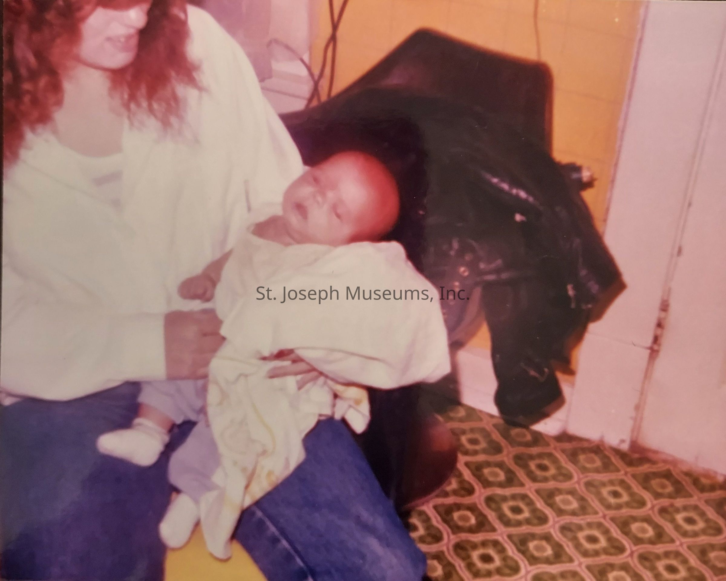 In this image, there is a woman with red hair sitting on a chair in a room with a patterned green and white floor, holding a sleeping baby wrapped in a light-colored blanket. The woman, wearing a white shirt and jeans, appears to be looking down at the infant with a gentle expression. Behind them, a black jacket is draped over another chair against a wall painted yellow, adding a cozy, intimate atmosphere to the scene. The image portrays a tender and peaceful moment shared between the woman and the baby.