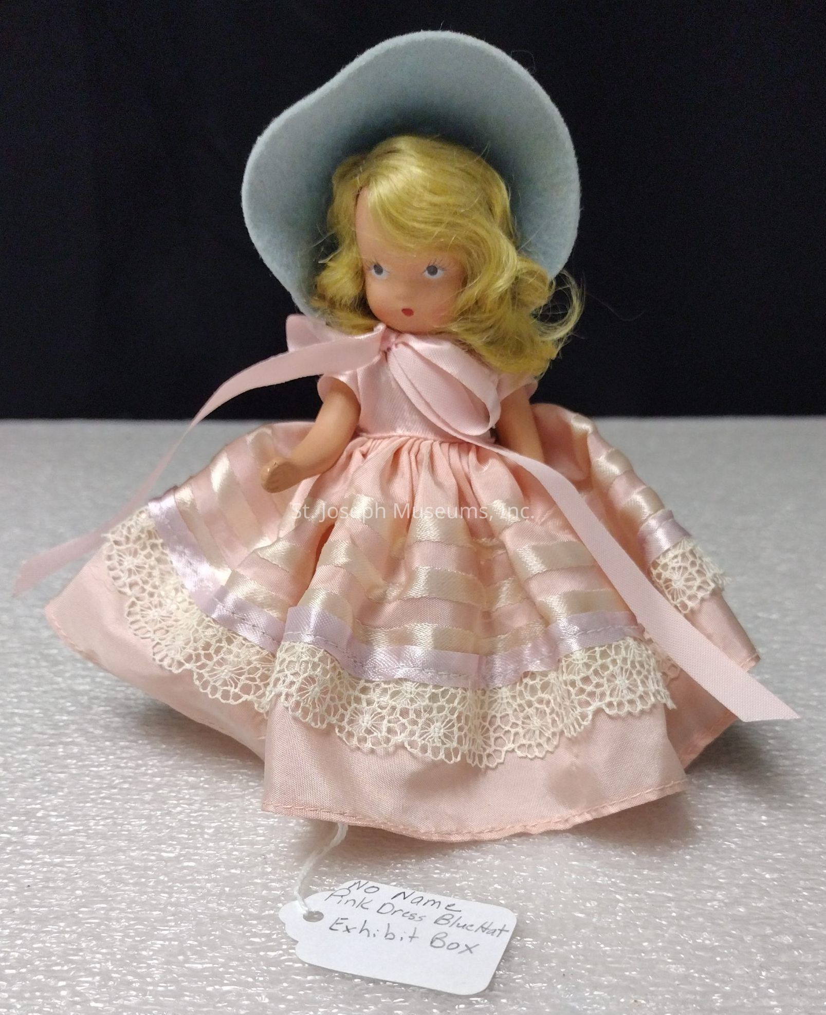 The image depicts a vintage-style doll with blonde hair wearing a light blue bonnet and an elegant pink dress that features lace and ribbon details. The attire evokes a classic and delicate aesthetic, reminiscent of bygone eras. The doll is positioned against a neutral backdrop, which highlights its intricate clothing details. A white tag attached to the doll contains handwritten words, likely identifying characteristics such as "No Name," "Pink Dress," "Blue Hat," and "Exhibit Box." This tag suggests the doll might be part of a collection or display. The craftsmanship of the doll, combined with its attire, gives it a charming and nostalgic appeal, conveying a sense of timelessness and tradition. The doll's expression is understated and serene, adding to its overall genteel and refined appearance. The image captures the miniature artistry and attention to detail which make collectible dolls cherished by enthusiasts. With the emphasis placed on the wardrobe's texture and color, the doll serves as a quaint embodiment of elegance and grace.
