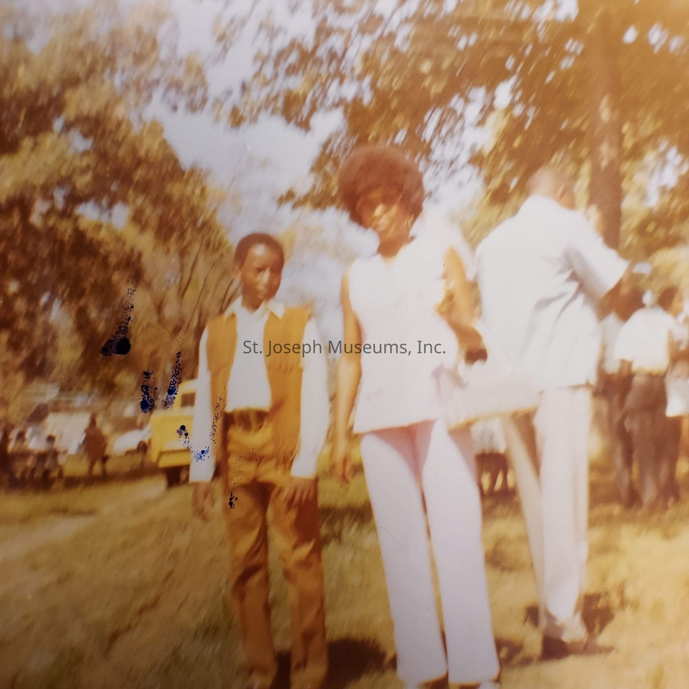 The image portrays a nostalgic, slightly faded scene, featuring a young boy and a woman walking side by side in an outdoor setting with tall trees and bright sunlight. The boy is dressed in a brown vest and trousers over a white shirt, looking calmly ahead. Beside him, the woman, with an Afro hairstyle, wears a sleeveless white blouse and matching pants, carrying a handbag in her left hand. The background is lively, with people socializing and a yellow vehicle parked nearby, evoking a warm, serene atmosphere of a casual outdoor gathering, possibly from decades past, as suggested by the vintage appearance and attire.