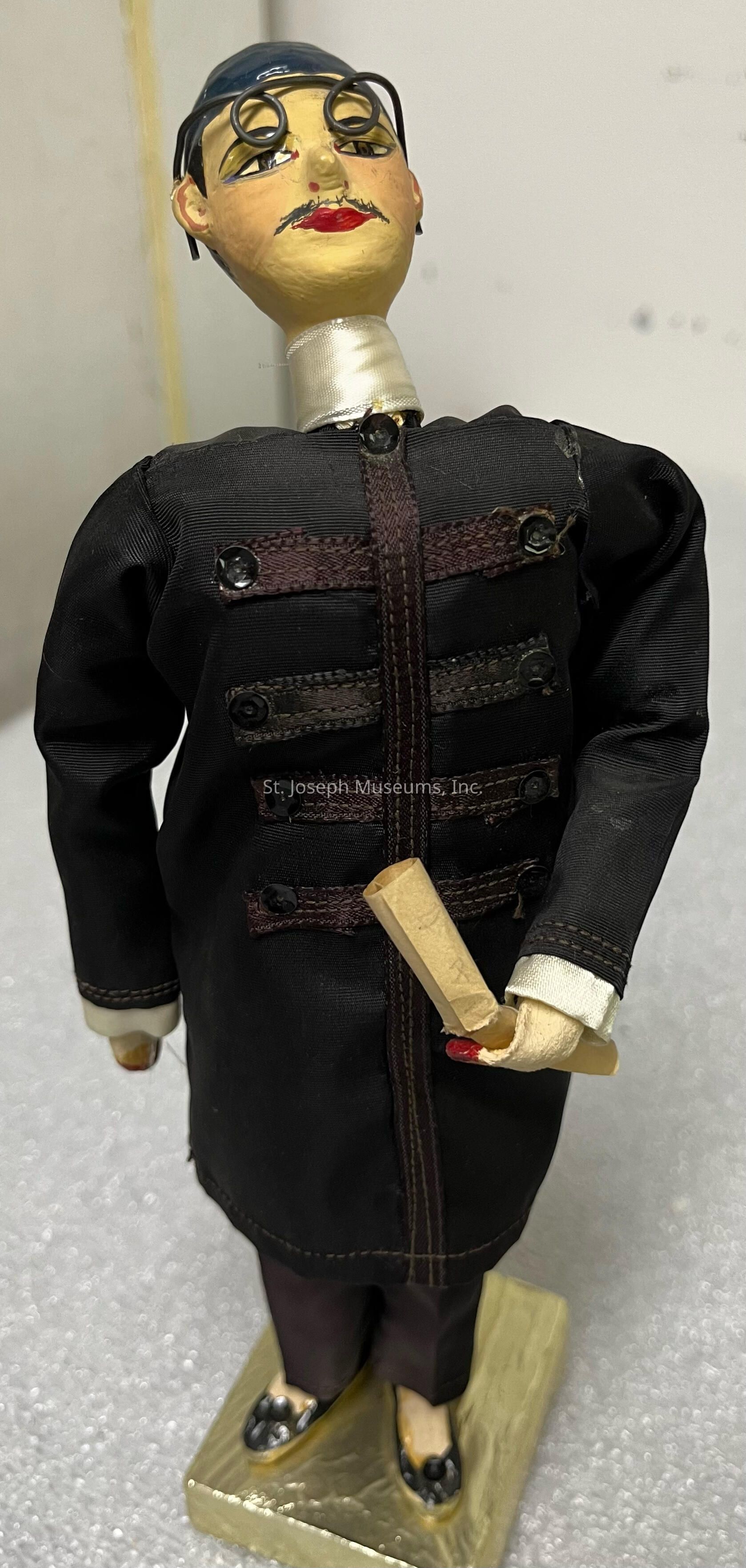 The image depicts a detailed puppet or figurine dressed in a black attire resembling a uniform adorned with decorative buttons and stitching. The figure stands upright on a textured rectangular platform and holds a rolled piece of paper in one hand. It has a uniquely crafted face with distinct painted features including red lips and a small mustache, while the eyes are accentuated by circular spectacles. The head is topped with a dark cap or hairpiece, and the neck is wrapped in a light-colored material. The overall craftsmanship suggests an artistic or theatrical theme, possibly representing a specific character or cultural style.