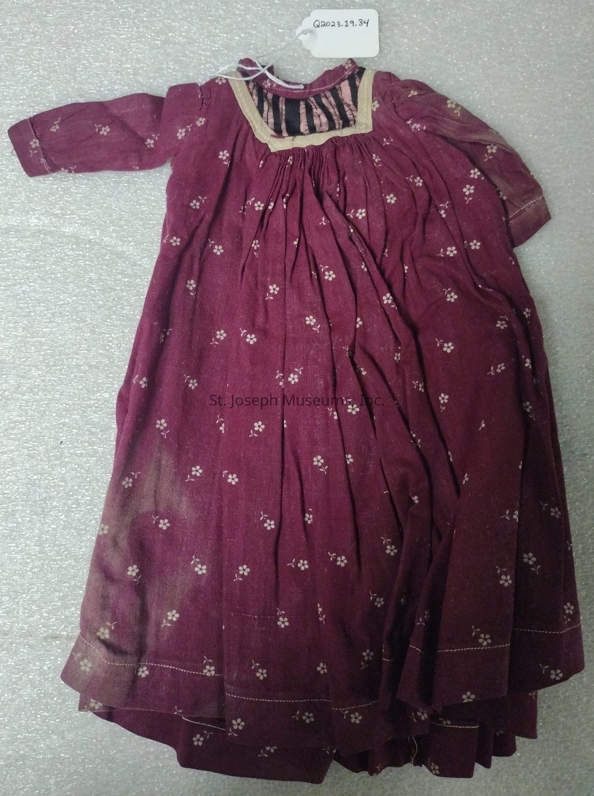 The image depicts a vintage child's dress, crafted from a rich maroon fabric adorned with a pattern of small, evenly spaced white flowers. The dress is long-sleeved and features a modest neckline, edged with a lighter trim that adds contrast to the overall deep hue of the garment. The fabric is gently gathered at the neckline, contributing to a voluminous skirt that suggests the ability to flow gracefully. A tag labeled "Q2023.19.34" is attached to the dress, possibly indicating a cataloging system for collections. The dress rests on a textured surface, enhancing the perceived antiquity and charm of this historical piece.