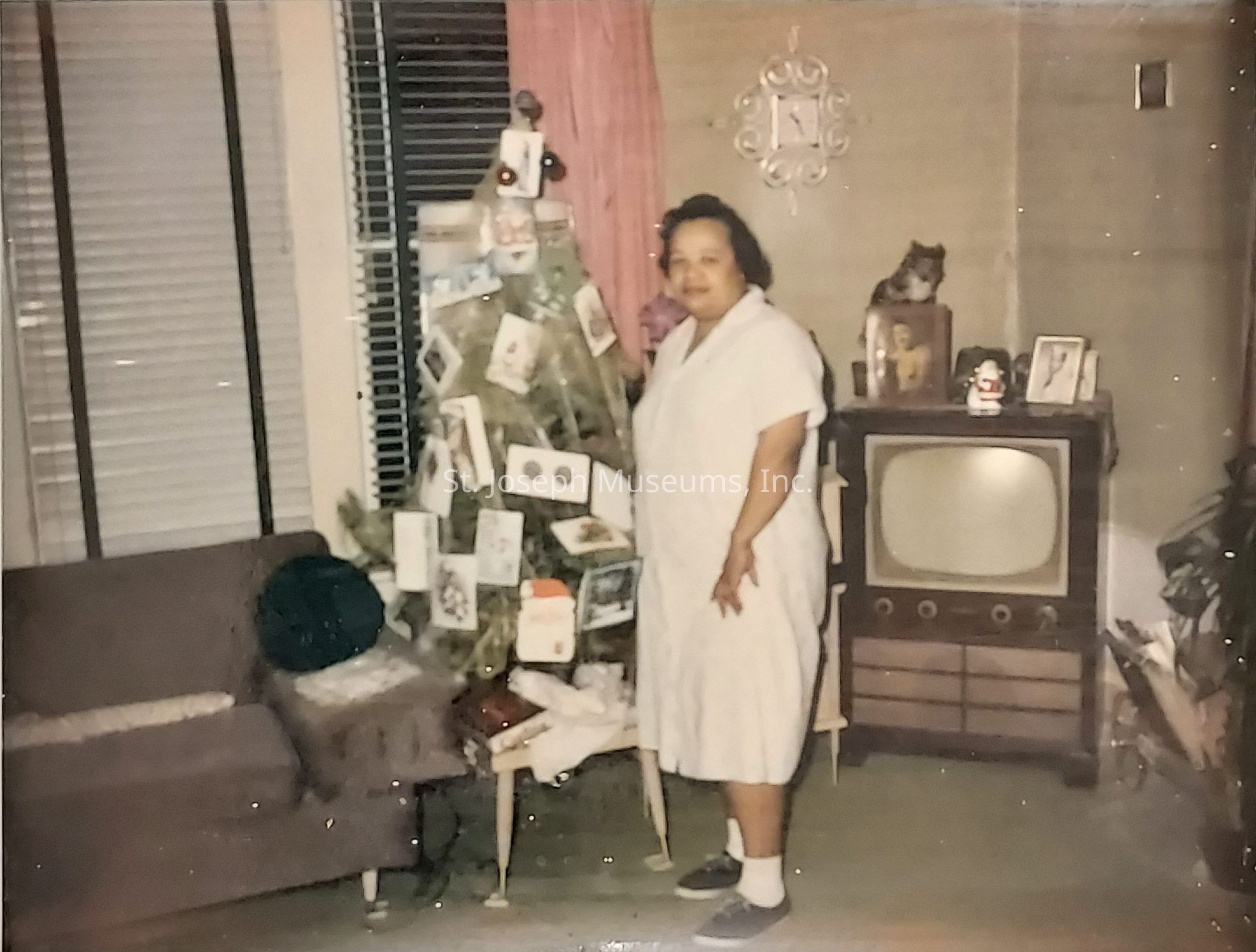 The image shows a vintage living room where a woman stands beside a small Christmas tree decorated with numerous greeting cards instead of typical ornaments. The tree is placed on a chair near a window with blinds, and a couch with a cushion is situated close by. To the right, an old television set, with knobs below the screen, sits on a cabinet. On top of the cabinet, there are framed photos and a small decorative piece, with a clock hanging on the wall above. The room has a cozy, nostalgic feel, suggesting a scene from many decades ago.