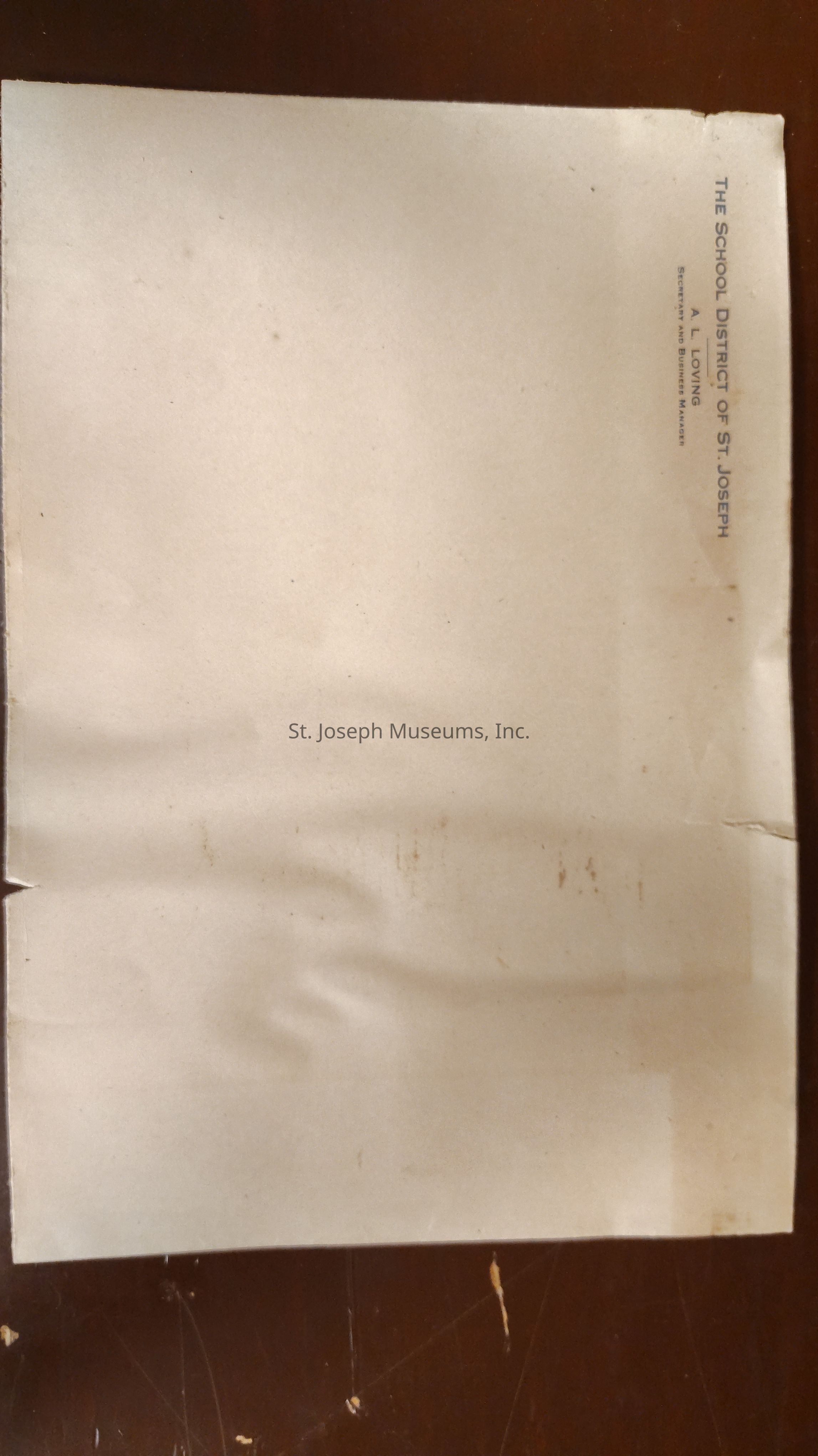 The image depicts a worn and partially marked sheet of paper featuring printed text in the upper right that reads "THE SCHOOL DISTRICT OF ST. JOSEPH" with additional lines identifying "A. L. LOVING" as "Secretary and Business Manager", showing signs of aging with visible creases and spots, suggesting it may once have been part of a larger document or served an administrative purpose in an educational context.
