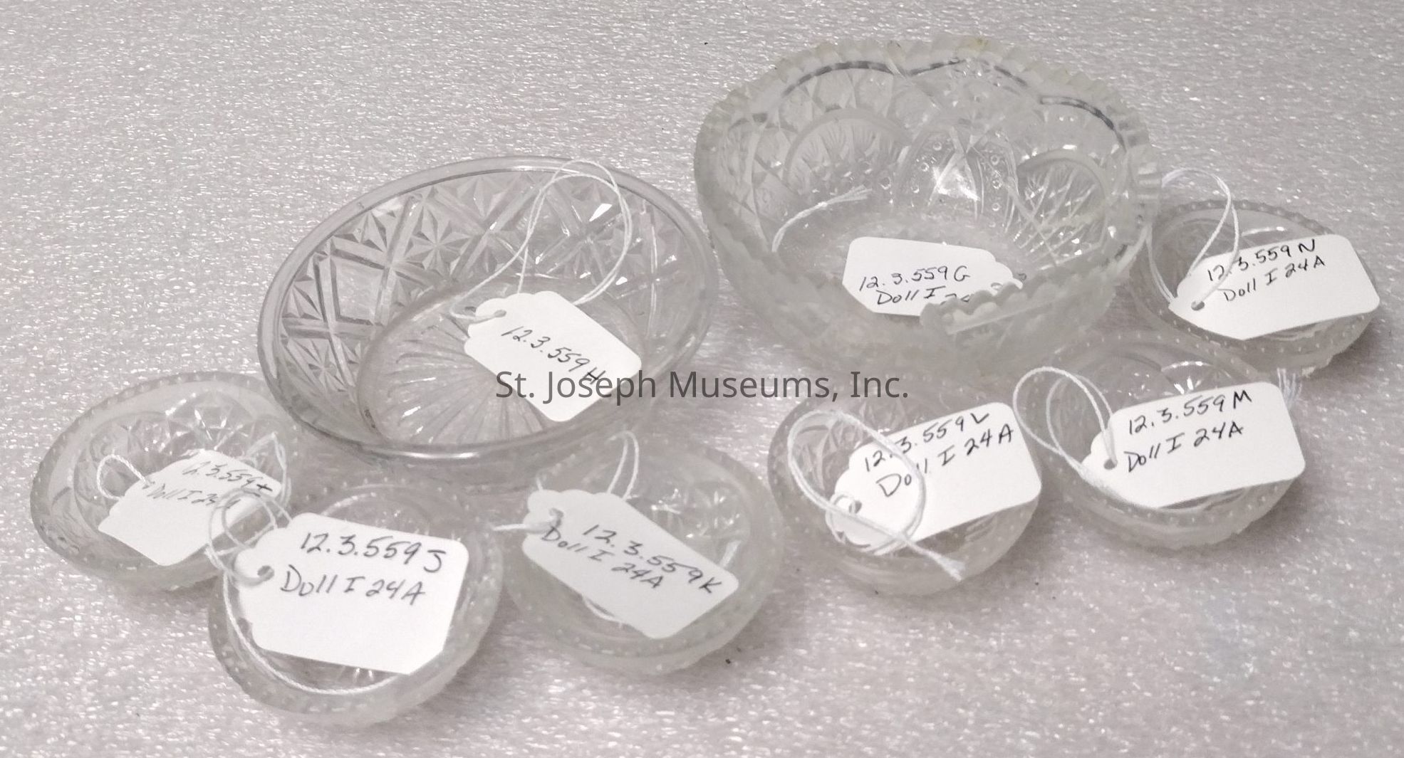 The image depicts a collection of intricately designed glass bowls and dishes, each adorned with distinct patterns and textures such as geometric shapes and floral motifs. These pieces are displayed on a textured white surface, and each one has a white tag attached with handwritten numbers and letters, possibly indicating catalog or inventory information. The tags suggest an organized presentation, likely for classification or sale purposes. The glass items vary in size, and their transparent nature allows the underlying design to be visible through the layers. The placement of the bowls and dishes is arranged in a manner that highlights their craftsmanship and delicate detailing.