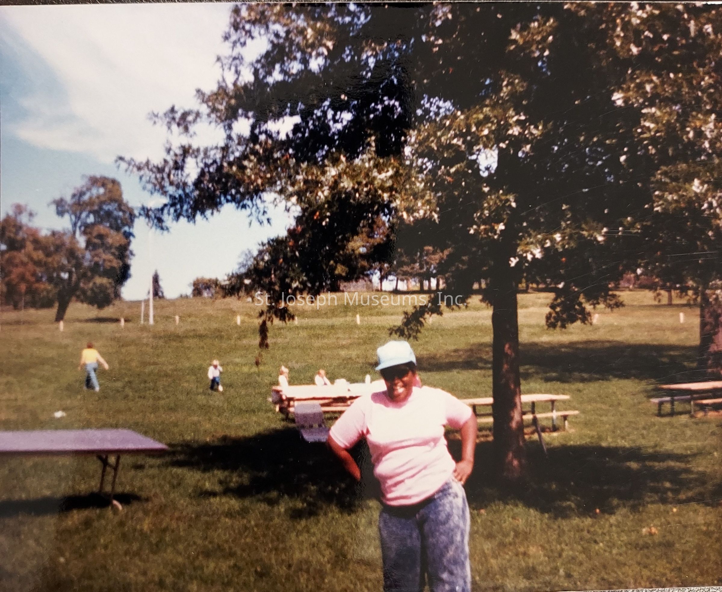 In this image, a person is standing in a sunny park with a relaxed and content pose, wearing a light-colored hat, a pink shirt, and blue jeans. The background showcases a sprawling green lawn dotted with a few people in the distance, engaged in leisurely activities such as walking or sitting at picnic tables. Several large, leafy trees provide shady spots throughout the area, casting soft shadows on the grass, and contributing to the tranquil and pleasant atmosphere. The sky is partly cloudy, with patches of blue visible, suggesting it's a mild, pleasant day ideal for outdoor activities. The person in the foreground appears to be enjoying the day, embodying a sense of joy and ease, with hands on hips, exuding confidence and a friendly demeanor. This scene captures a quintessential outdoor experience, combining elements of nature, relaxation, and community, reflective of a day spent connecting with nature and perhaps enjoying the company of friends or family in a scenic and serene setting.