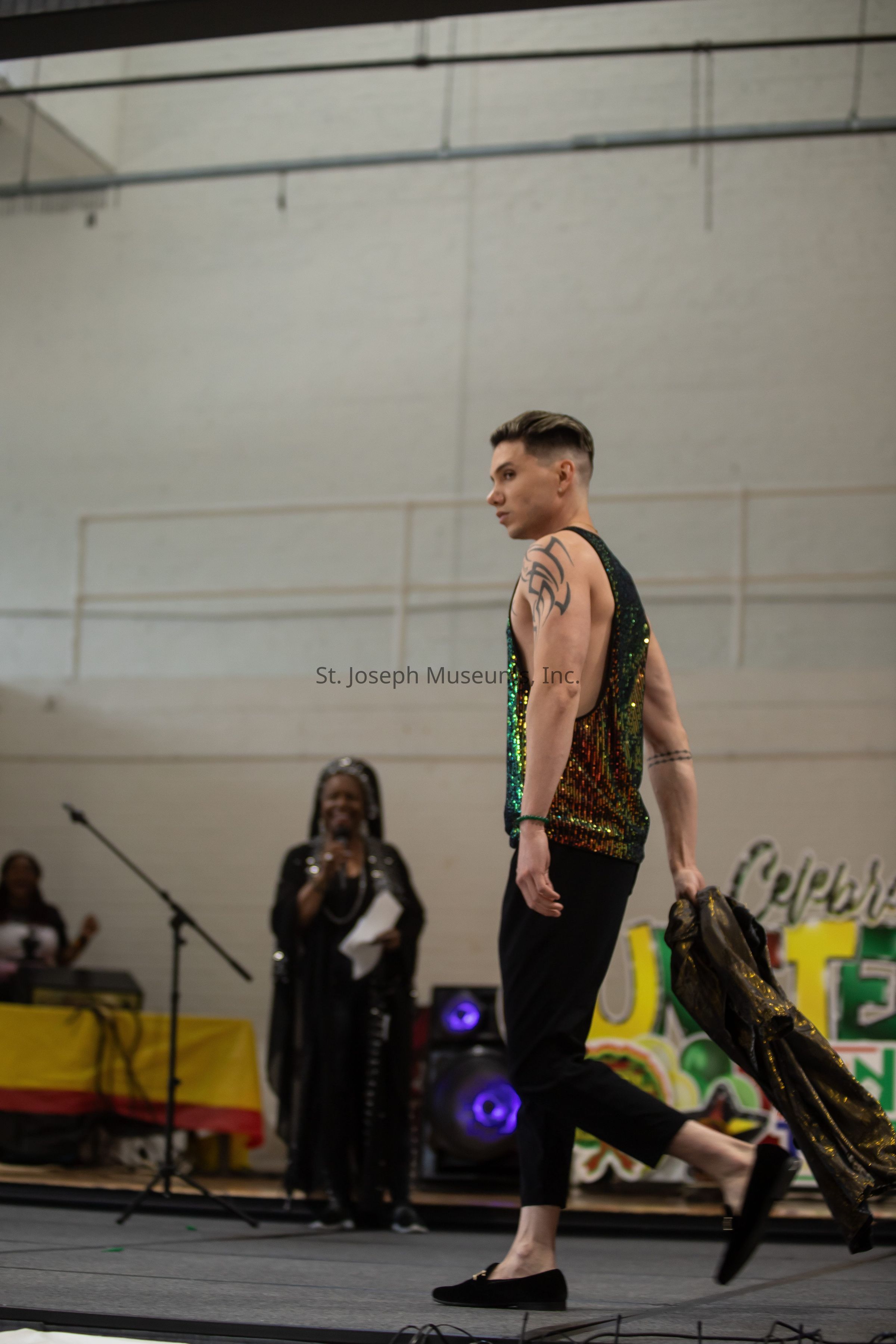 In the image, a person with short hair is striding confidently down a runway, wearing a striking, shimmering sleeveless top paired with black pants and shoes, while holding a jacket casually in one hand. A tattoo is visible on their arm, enhancing the bold and trendy appearance. Behind them, an individual with a microphone stands against the backdrop of colorful speakers and a vibrant sign, possibly indicating an event or celebration. The setting appears to be indoors with a casual ambiance, as suggested by the high ceiling and visible structural elements like beams and wires.