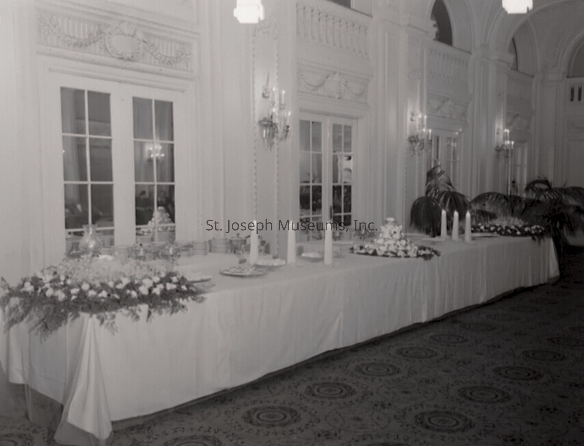 A black-and-white photo of the northwest corner of the St. Joseph, Missouri Robidoux Hotel's Crystal Room, displaying a set-up banquet table draped in a white cloth, adorned with floral arrangements, tall candles, and elegantly arranged tableware, all set against the backdrop of grand French doors and intricate wall moldings.