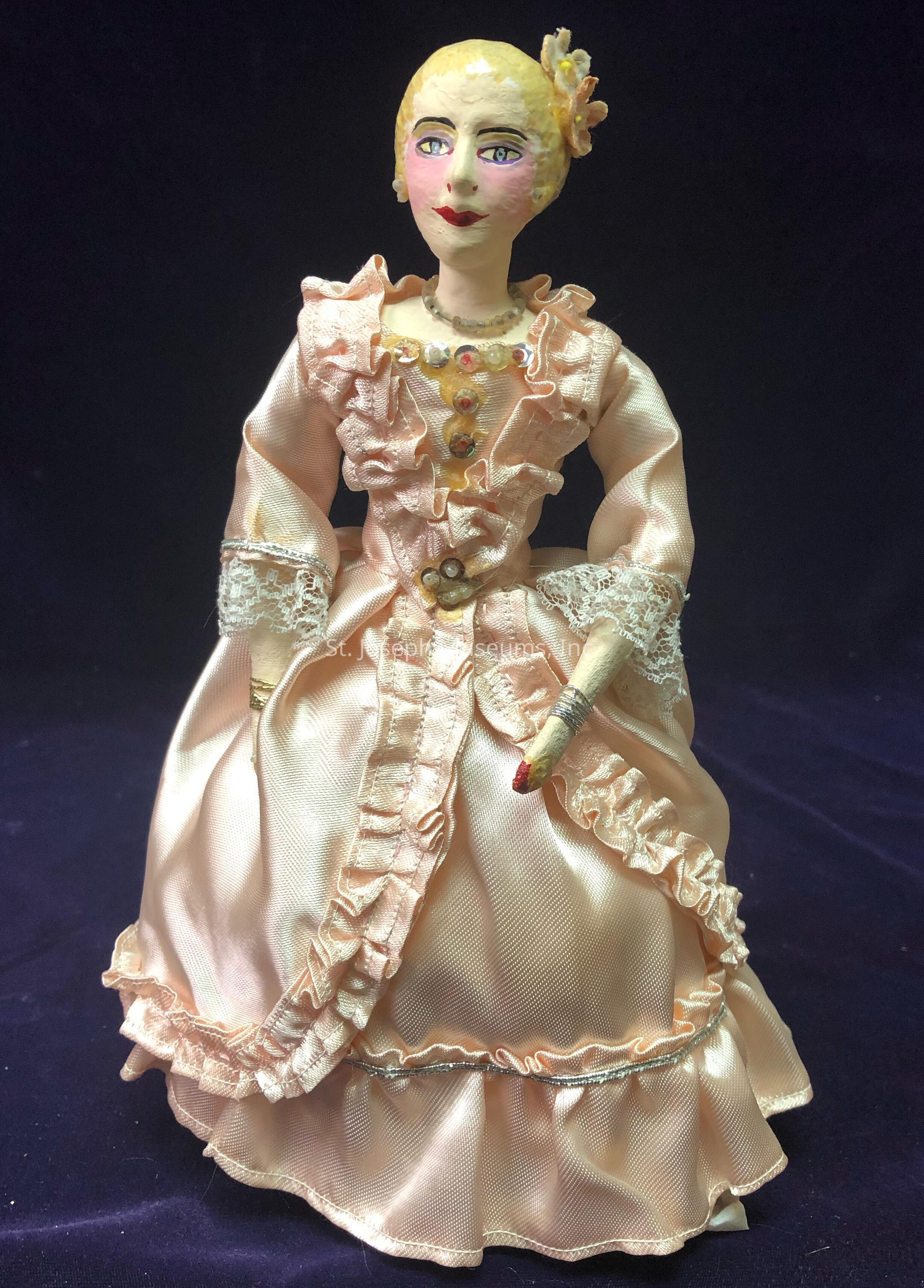 The image depicts a handcrafted doll dressed in an elegant, pastel pink gown adorned with ruffles and lace details, exuding a vintage charm. The gown features a fitted bodice embellished with decorative beads, while the sleeves are gracefully adorned with lace trim. The doll's face is carefully painted, showcasing rosy cheeks, defined eyebrows, and lips tinted in a rich red hue, conveying a classical and sophisticated appearance. Atop its head, a stylish hairstyle is accented with a small floral accessory, complementing the overall aesthetic of the attire. The doll’s hands, crafted with attention to detail, add to the lifelike quality of the figure. The dark, plain background enhances the contrast and highlights the intricate craftsmanship and delicate fabric of the doll's attire, making the doll appear almost lifelike. This exquisitely crafted figure embodies an artistic representation of a bygone era, capturing the viewer’s attention with its detailed and thoughtful design.