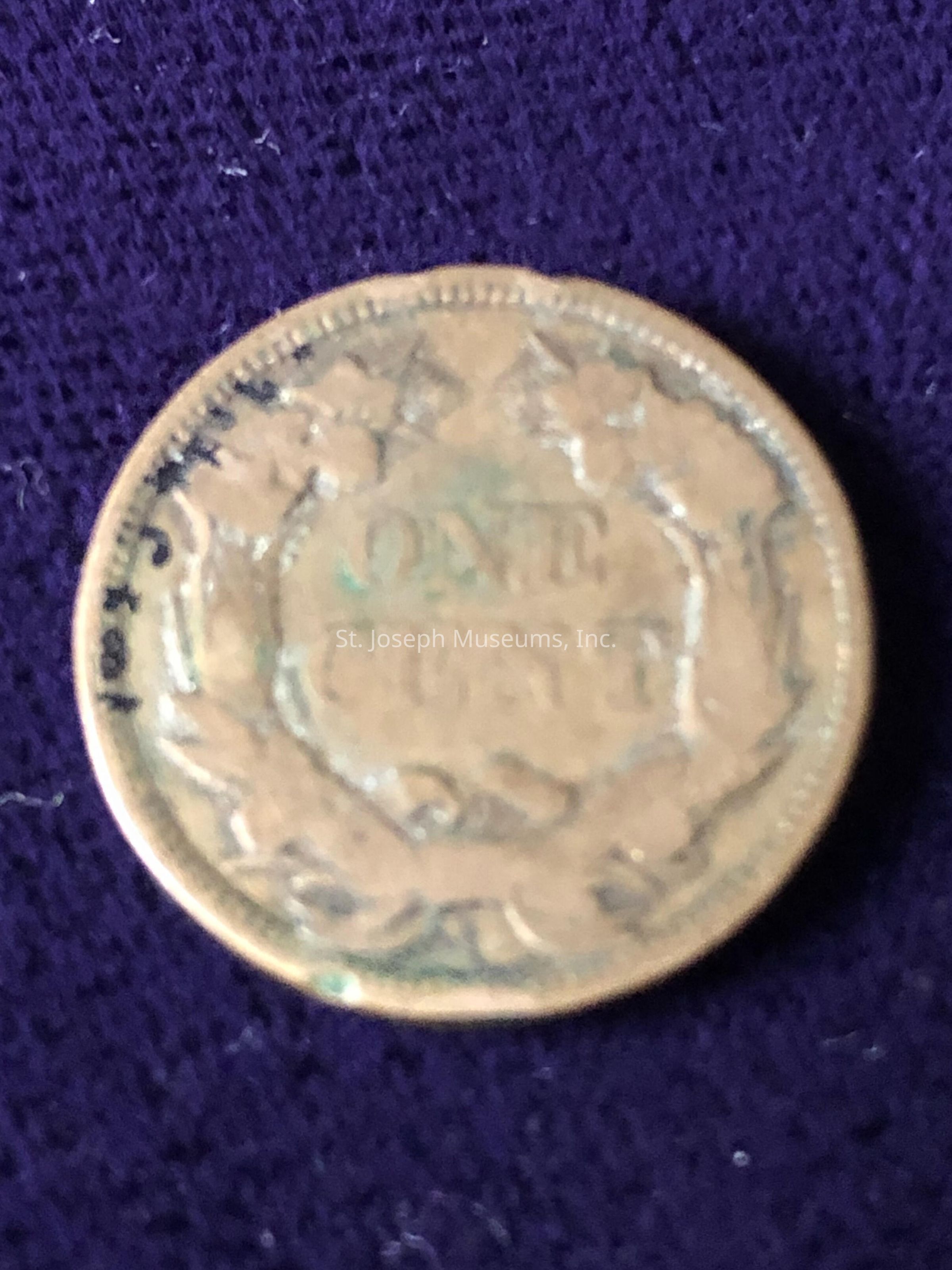 The coin, showing considerable wear and age, features the inscription “ONE CENT” prominently in the center, surrounded by a wreath design. Its edges show signs of discoloration and patina, which add to its antique appearance. The intricate detailing of the wreath is partially obscured due to the coin's worn condition, indicating many years in circulation. The fabric background provides a contrasting backdrop, highlighting the coin's coppery hue with subtle greenish tarnish that suggests its historical significance.
