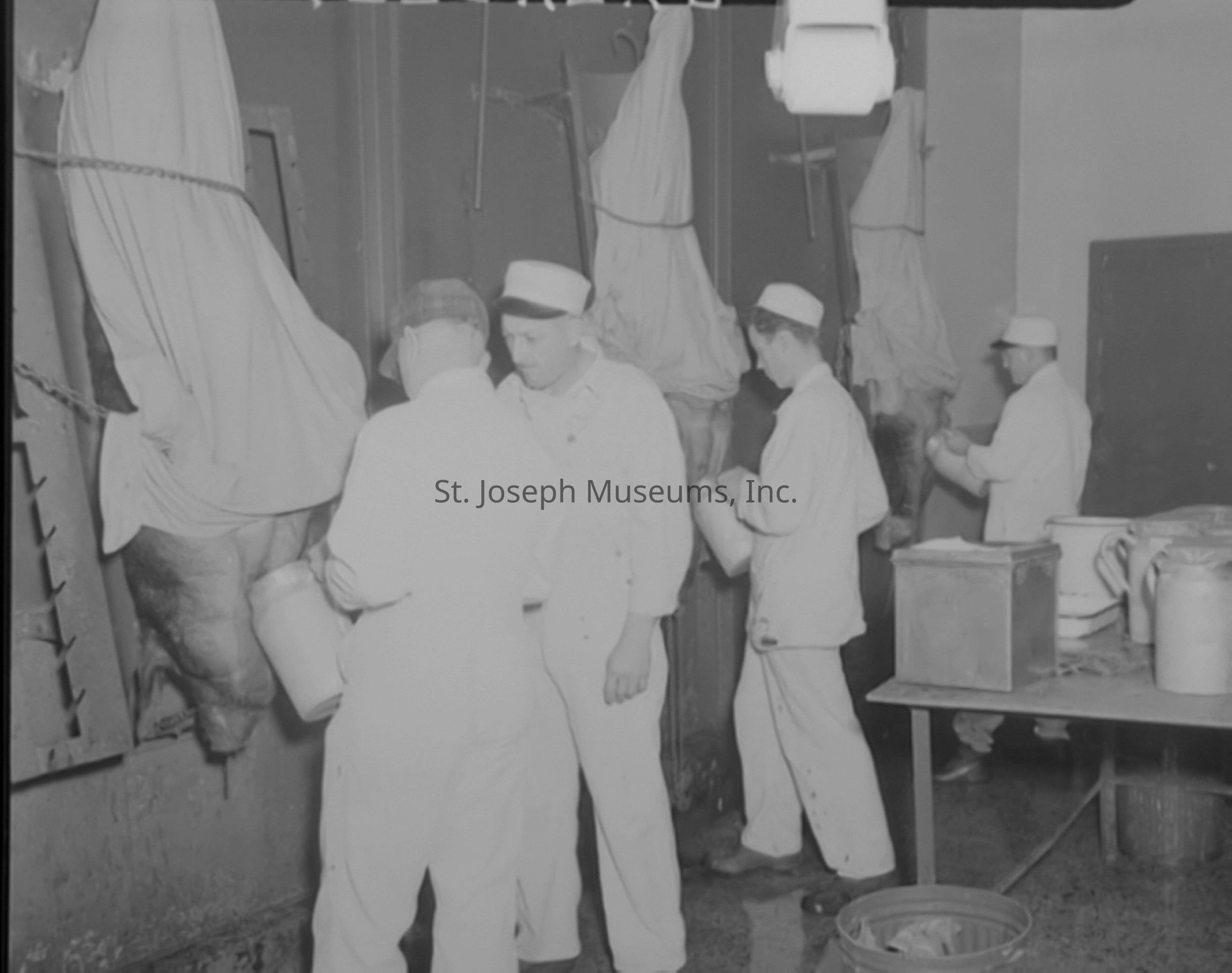 Black-and-white photograph of the Anchor Serum Company facility on Lower Lake Road in St. Joseph, Missouri. Several workers in white uniforms and caps are processing animal carcasses; they are engaged in various tasks, including handling large metal containers, in an industrial setting with hanging carcasses covered by sheets.