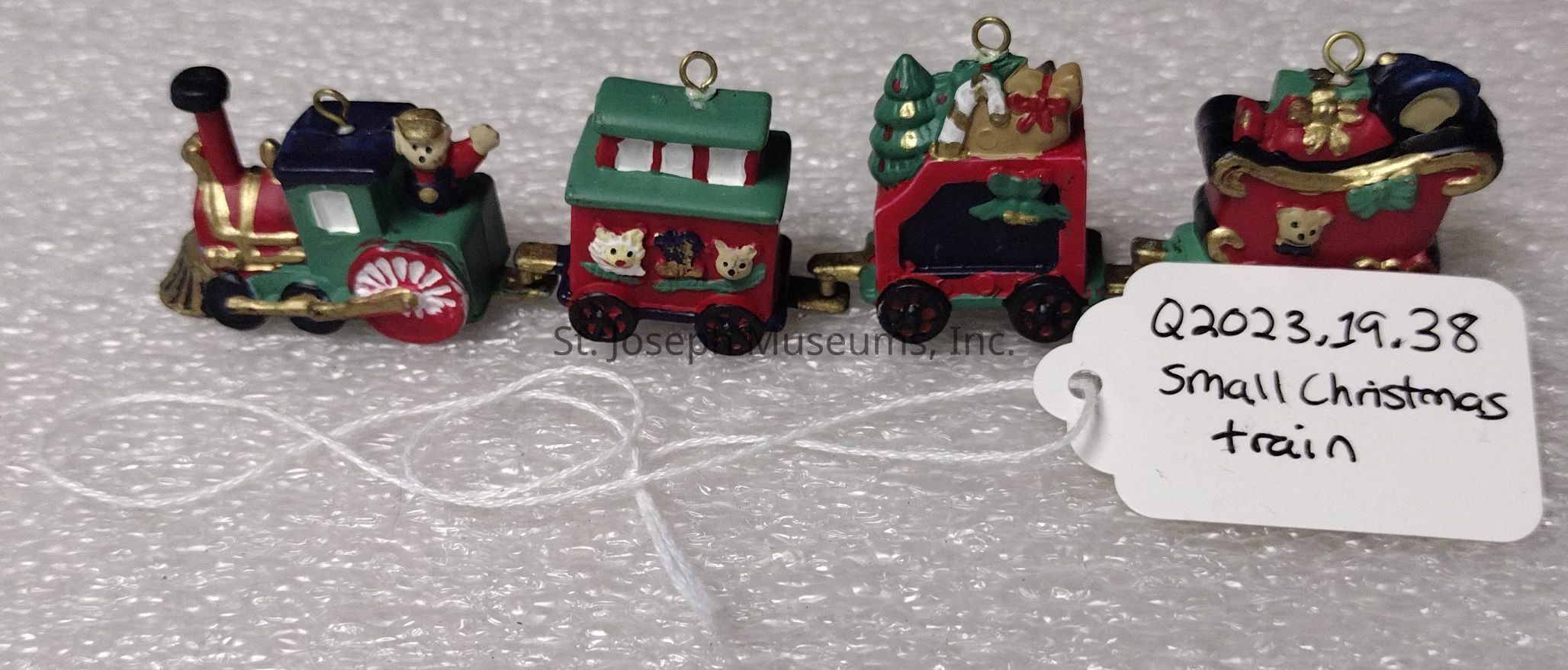 The image depicts a small decorative Christmas train consisting of four colorful carriages in a festive design. The train is adorned with vibrant holiday-themed elements, including ornaments, trees, and figures like teddy bears, painted in cheerful red, green, gold, and white. Each carriage has a unique design, with the first carriage featuring a locomotive and a little bear conductor. The second carriage has intricate animal figures in the windows, the third is loaded with gifts and a Christmas tree, and the fourth resembles a sleigh with presents. The entire train rests on a textured white surface, and a tag is attached with the inscription "Q2023, 19, 38 Small Christmas train," suggesting it might be an exhibit or collectible item.