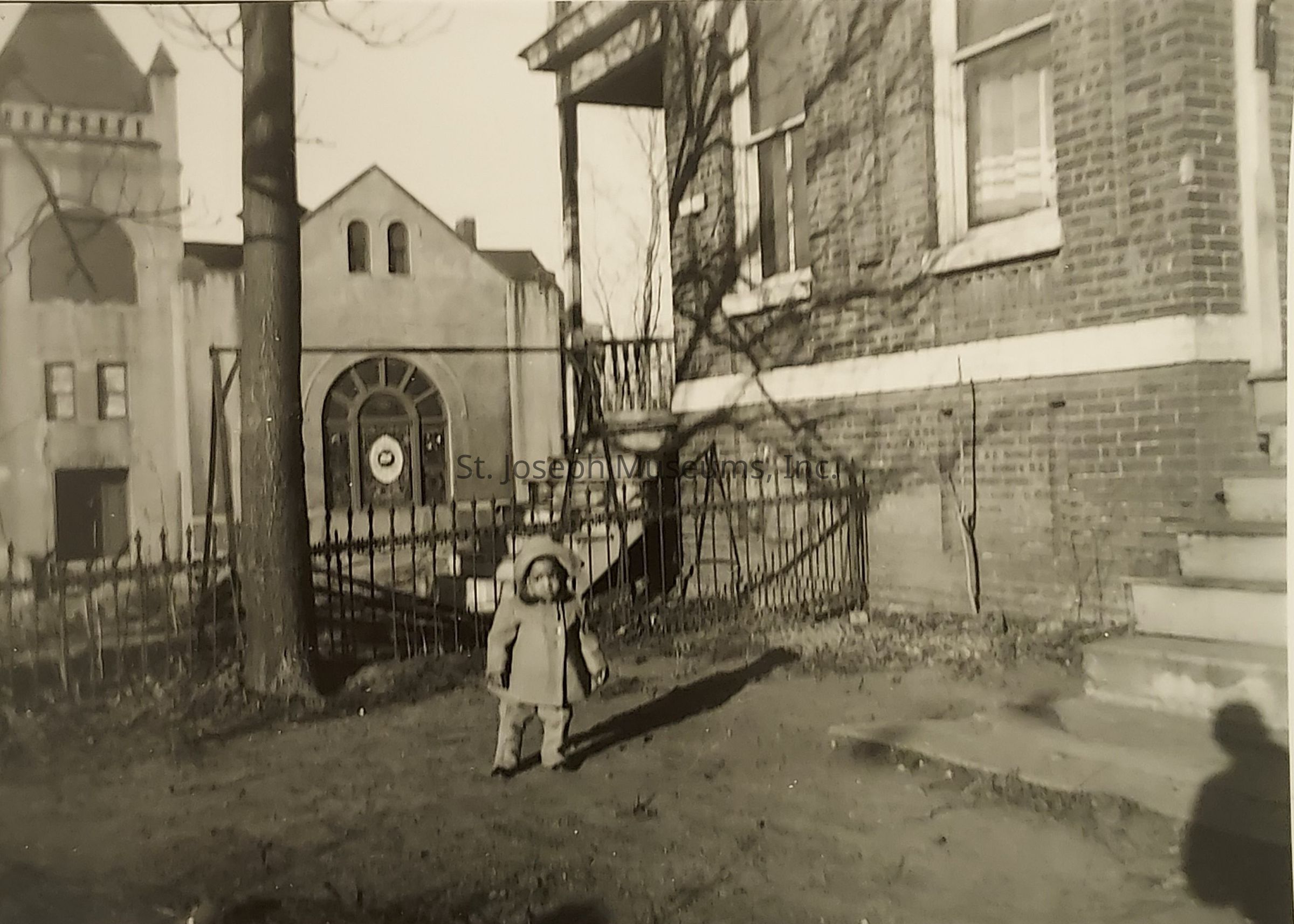 In a sepia-toned image reminiscent of the past, a small child stands in the foreground wearing a coat and hat, casting a long shadow on the ground. To the right, a brick building with a window and staircase provides a backdrop, while a wrought iron fence adds to the nostalgic atmosphere. Across a narrow passageway, another building features arched windows and a circular detail above the entrance, suggesting architectural interest typical of the early to mid-20th century. Bare trees, devoid of leaves, indicate a chilly season, enriching the historical mood.
