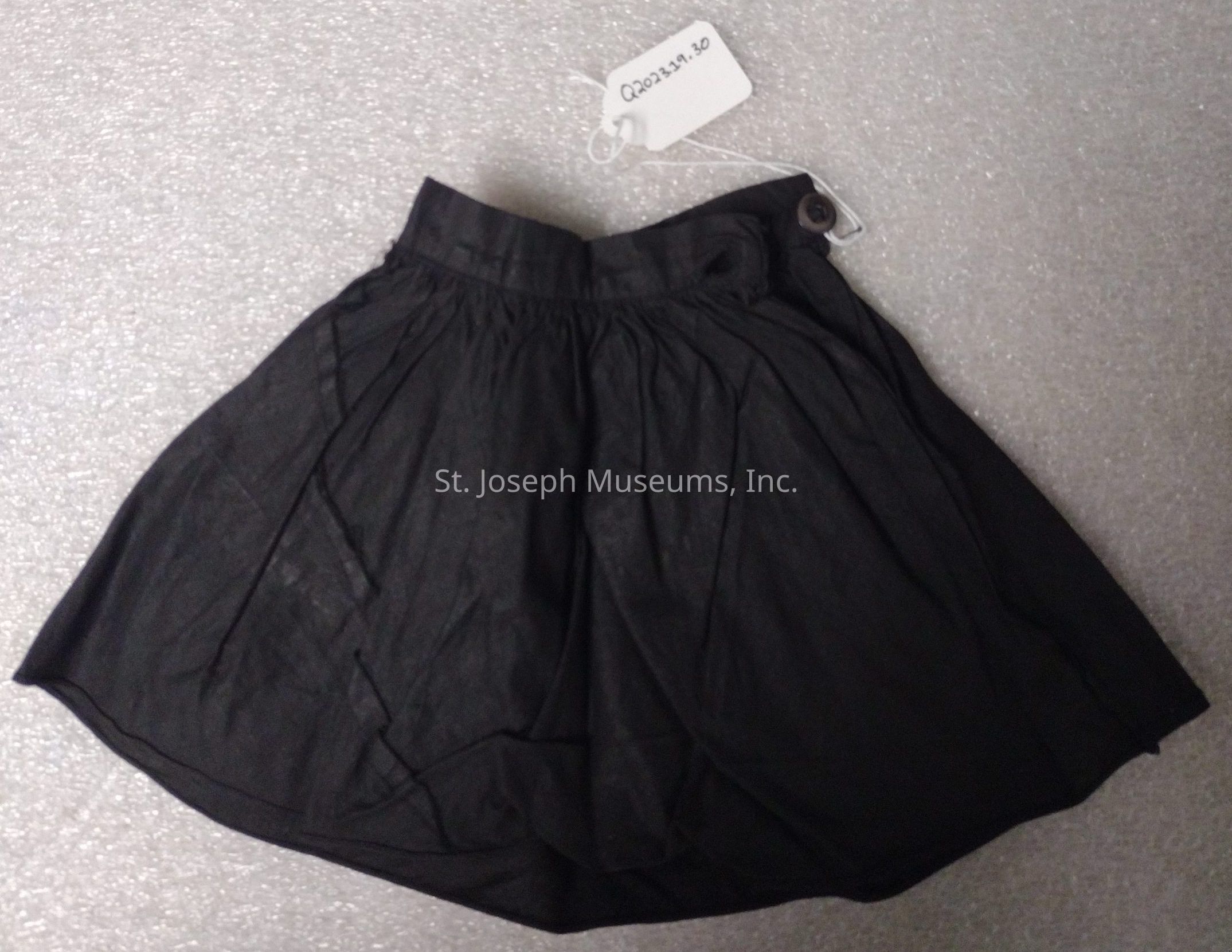 The image features a pair of neatly arranged black shorts made from a lightweight, sheer fabric that drapes softly, with visible pleats creating a slightly flared appearance. The waistband is elastic, providing a comfortable fit for the wearer, and it has an extra button attached on one side which hints at a possible adjustable feature or decorative design. There is also a small white tag attached with a string, containing an alphanumeric code ("Q2021.20"), possibly for cataloging or identification purposes. The shorts are placed on a textured light gray surface that contrasts with the dark fabric, enhancing the visibility of the garment. Overall, the presentation suggests these shorts might be part of a museum collection or an archival catalog, given the attention to detail in tagging and arrangement.
