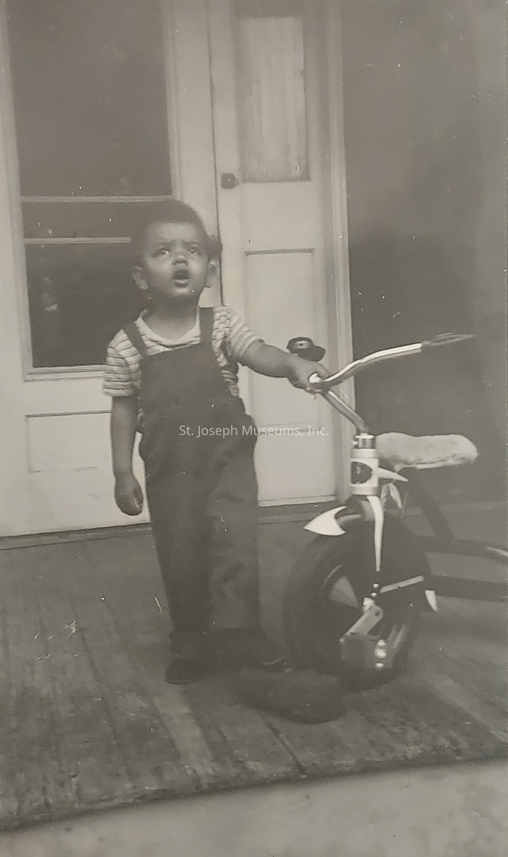 In this vintage black and white photograph, a young child is captured standing on a porch in front of a wooden door, wearing striped short-sleeves and dark overalls, while holding the handlebars of a tricycle with one hand; the child appears to be looking upwards with an expression of curiosity or surprise, and the setting hints at a candid and nostalgic moment, with the textures of the wooden floor and the house in the background contributing to the timeless atmosphere, conveying a sense of innocence and childhood exploration, especially as the cozy, slightly dim lighting adds a warm, sentimental touch to the scene, immortalizing a simple yet precious slice of everyday life.