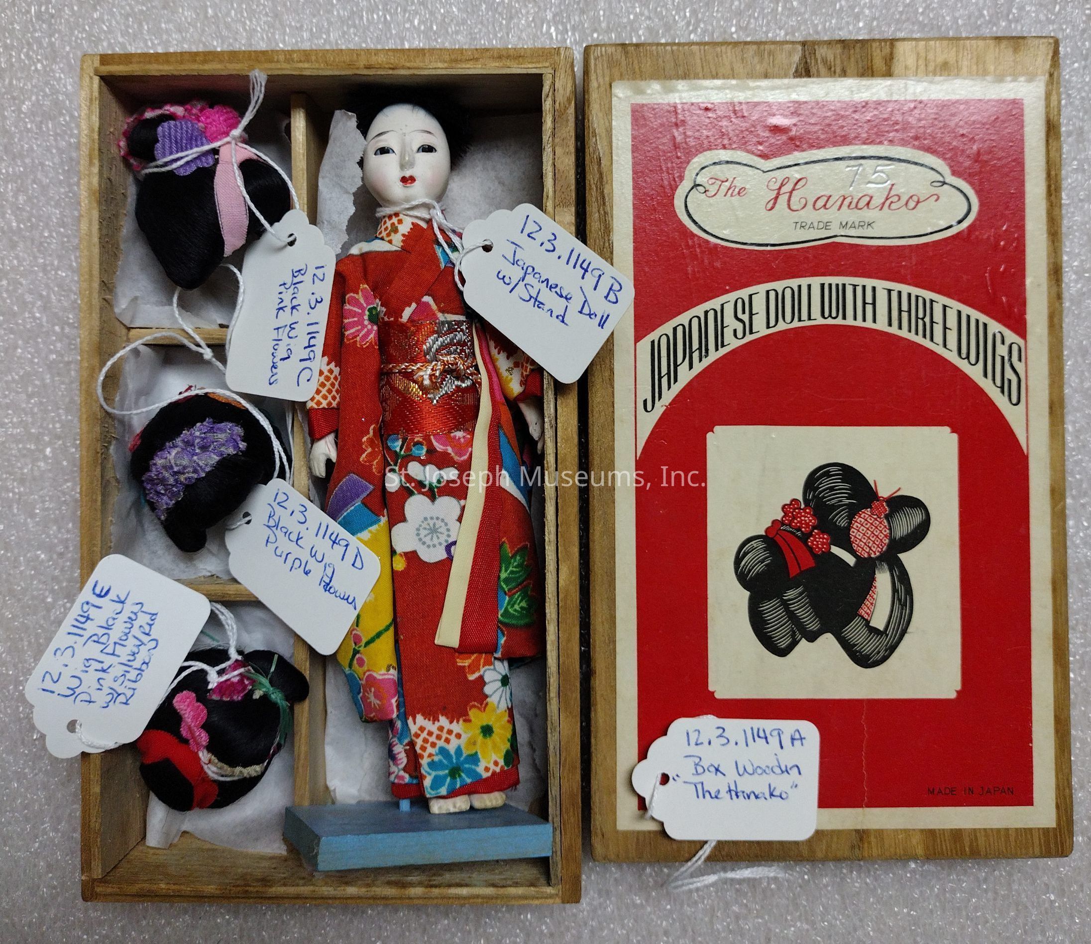 The image shows a traditional Japanese doll dressed in a vibrant red kimono adorned with floral patterns, housed in a wooden box. Accompanying the doll are three intricately crafted wigs, labeled and stored alongside it. Each wig label has distinct notes written in blue ink describing the color and style: black with pink flowers, black with purple flowers, and another style mentioned in similar detail. The packaging, which prominently features red and white colors, is labeled "The Hanako" and "Japanese Doll With Three Wigs," indicating that it is a product made in Japan. Decorative motifs and the box design reflect traditional Japanese aesthetics. The doll and accessories are carefully positioned and tagged, suggesting that they are part of a cataloging system or collection. The tags on each component include unique identifiers, detailing specifics of each item, such as the type of wig or the fact that the doll includes a stand. These tags suggest careful archival or curatorial practices, possibly for a museum or private collection.