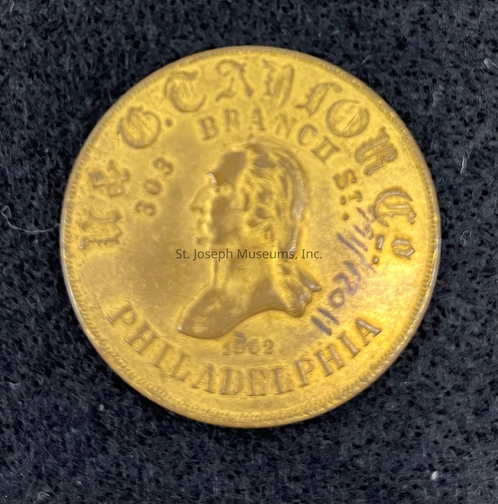 The image features a round, gold-toned token with an embossed profile of a person facing left at its center, framed by text that reads "W.J. Taylor 303 Branch St. Philadelphia" along the outer edge; this text suggests the token may be a type of trade or advertising piece from a business or organization, likely created in connection with the establishment's location in Philadelphia, and the surface of the token appears slightly worn, indicating some age or use, while the background is a textured, dark material that contrasts with the bright metallic sheen of the token, enhancing the clarity of the details and inscriptions.