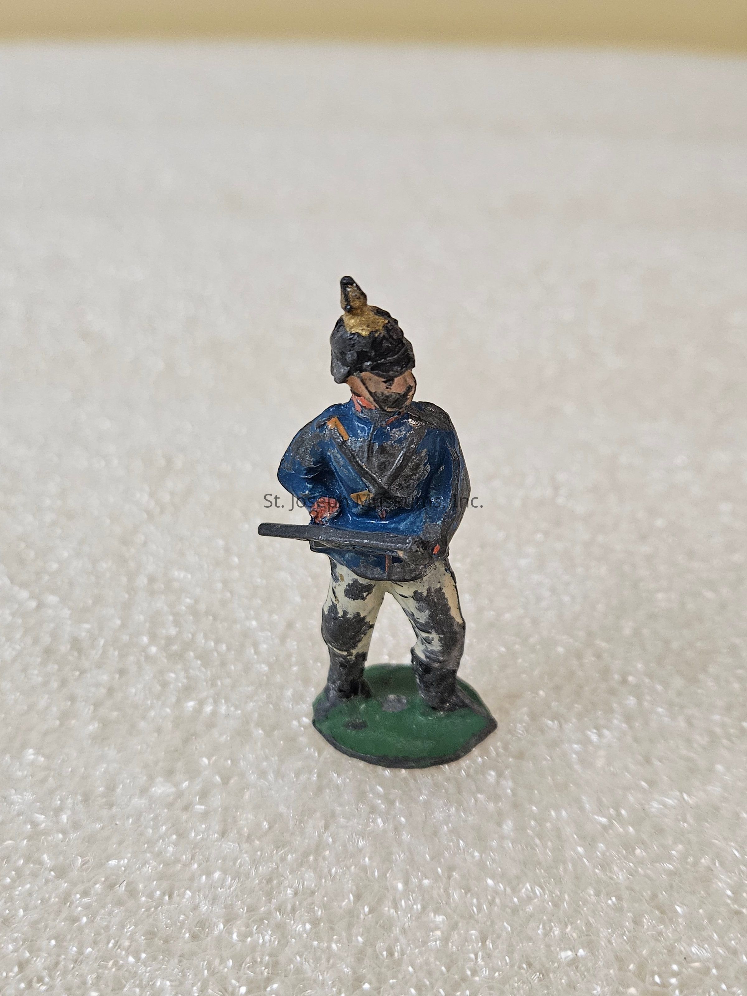 2024.5.11 A: The image depicts a small, intricately detailed figurine of a soldier standing on a textured surface, likely representing a historical or model military figure. This figurine is adorned in a blue uniform with contrasting dark and light details, featuring a prominent helmet with a plume on top, adding to its historical allure. The soldier is posed with a rifle held across the chest, exhibiting a stance of readiness or alertness. The small base upon which the figure stands is green, providing a slight elevation and stability. The meticulous paintwork on the figurine highlights the folds and contours of the uniform, giving the piece a sense of realism despite its diminutive scale. The textured surface beneath the figure contrasts with the smooth, defined lines of the soldier, accentuating and highlighting its presence, suggesting it might be part of a larger collection or diorama. The detailing and historical costume suggest it may be a piece collected by enthusiasts of military history or model figurines.