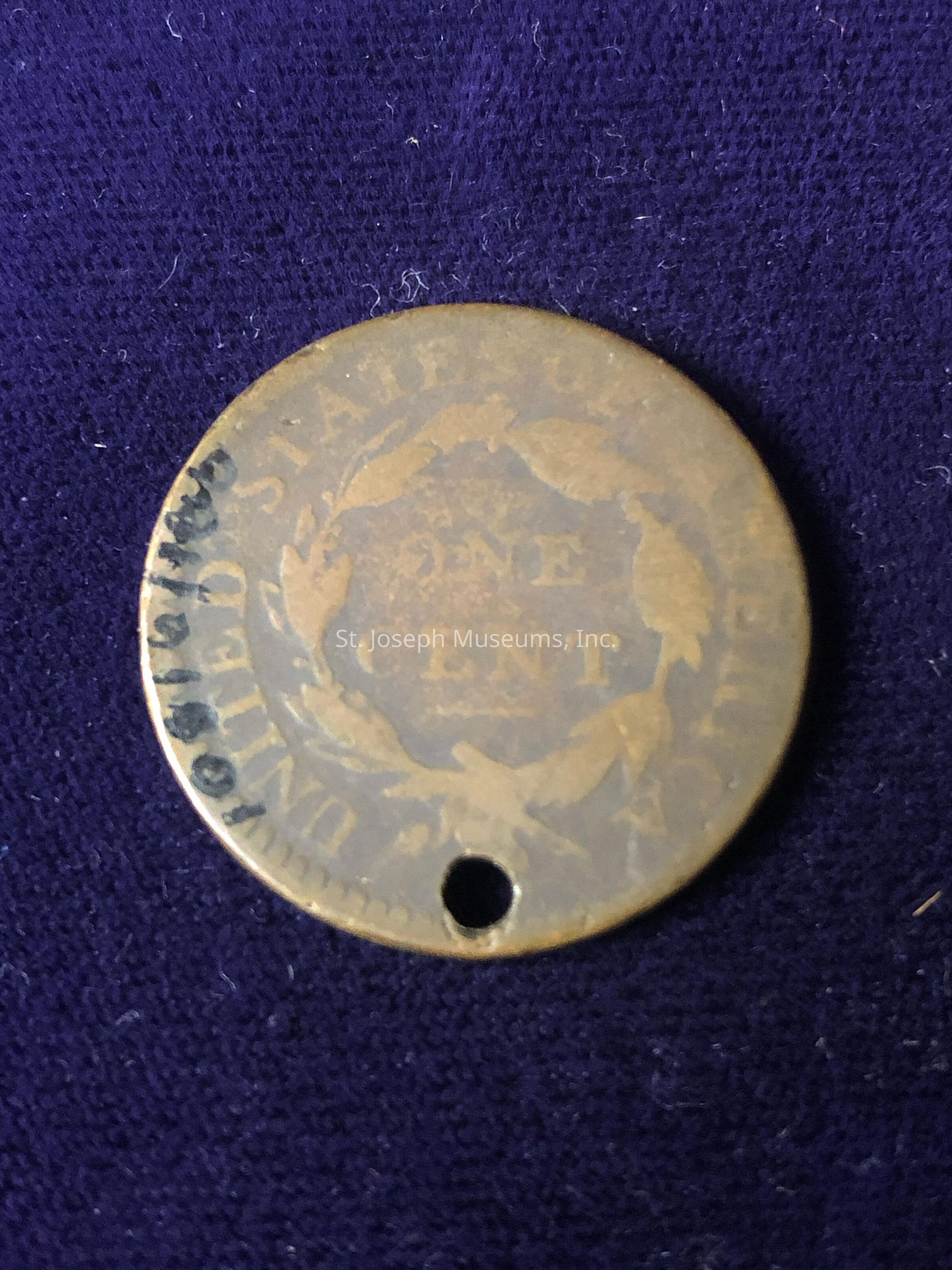 The image shows an old, worn-out coin with the words "ONE CENT" visible in the center, surrounded by a decorative wreath. The coin has a hole near the edge, possibly indicating it was once used as a pendant or for another purpose. The inscription around the wreath reads "UNITED STATES OF AMERICA." The coin rests on a dark blue surface, which provides a contrasting background that highlights the coin’s details. The coin's surface appears aged and tarnished, with some areas darker than others, indicating significant wear and the passage of time.