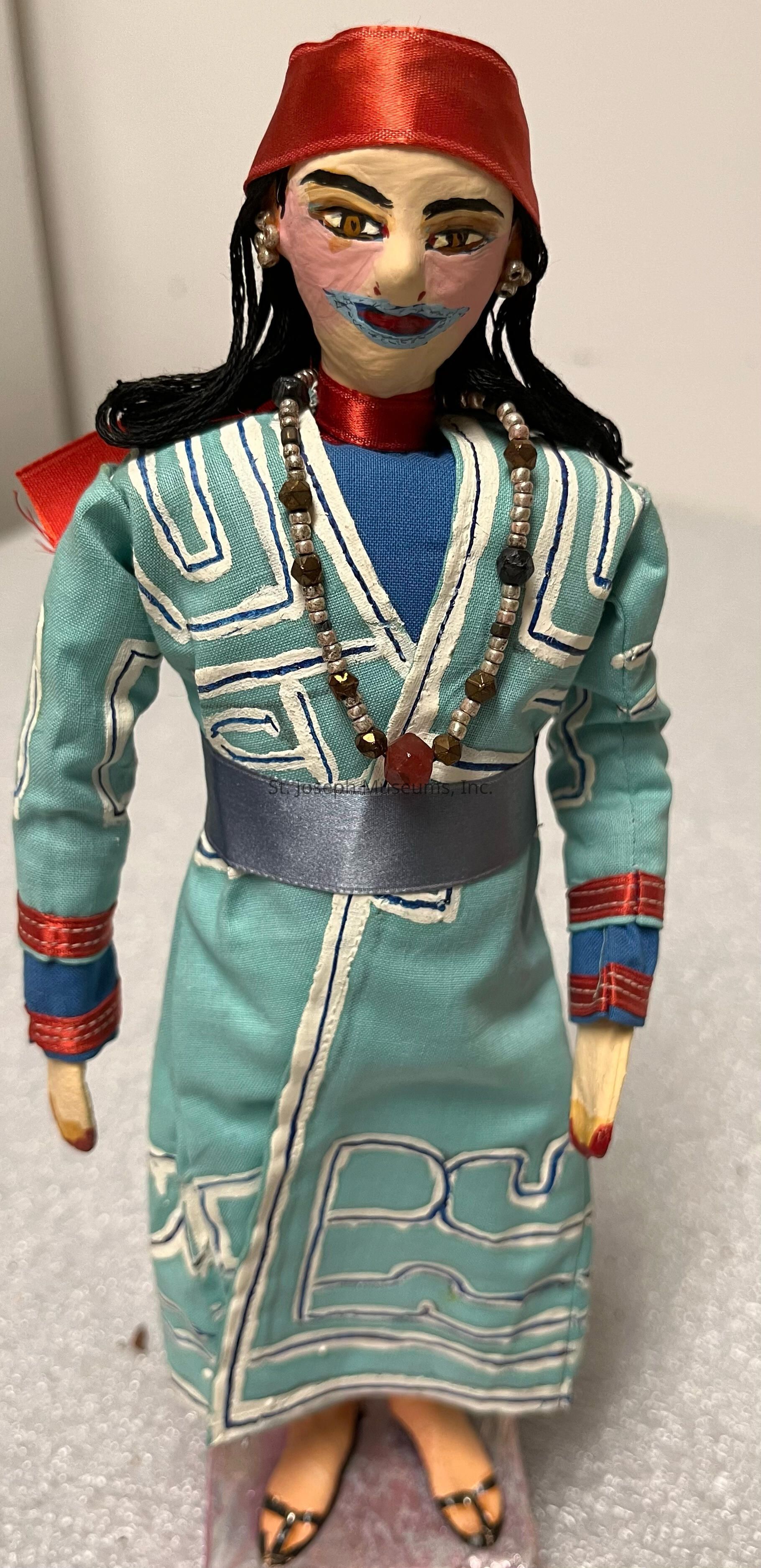The image depicts a handcrafted doll adorned in a vibrant turquoise outfit with white and blue embroidered patterns. The doll is wearing a red headscarf, which complements its striking facial features painted vividly, highlighting the eyes and lips. A pearl and bead necklace with a red accent stone adds a touch of elegance to its attire. The sleeves of the garment are detailed with red and blue bands, matching the ribbon around the doll's waist, and its black hair cascades down from under the headscarf. The craftsmanship is intricate, showcasing a combination of textile art and painting, capturing a cultural essence.