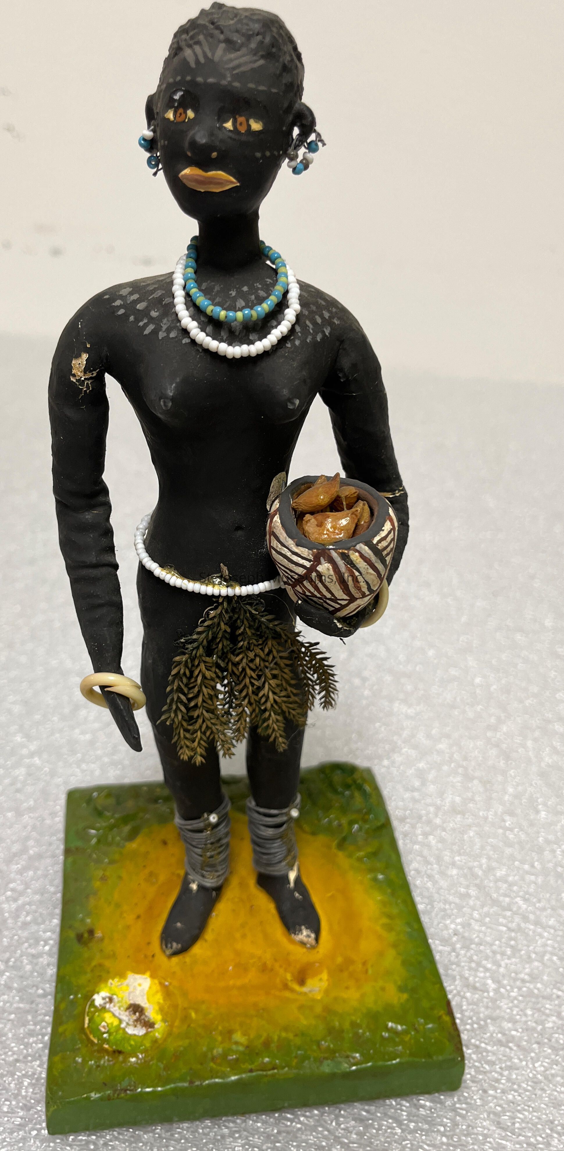 The image displays a detailed figurine of a person standing on a green and yellow base. This figure is adorned with various accessories, including white and blue beaded necklaces and earrings. The figurine has yellow-painted eyes and lips, adding contrast to the dark surface. It holds a bowl filled with a substance resembling nuts or seeds. The figure wears a textured skirt made of plant-like material and circular decorations. Its ankles and upper arms seem to be wrapped with coiled material, adding an intricate element to the overall appearance. The sculpture's design appears to symbolically reflect cultural or traditional attire, presenting an artistic representation characterized by both simplicity and attention to detail, conveying a sense of cultural heritage and artistic expression.