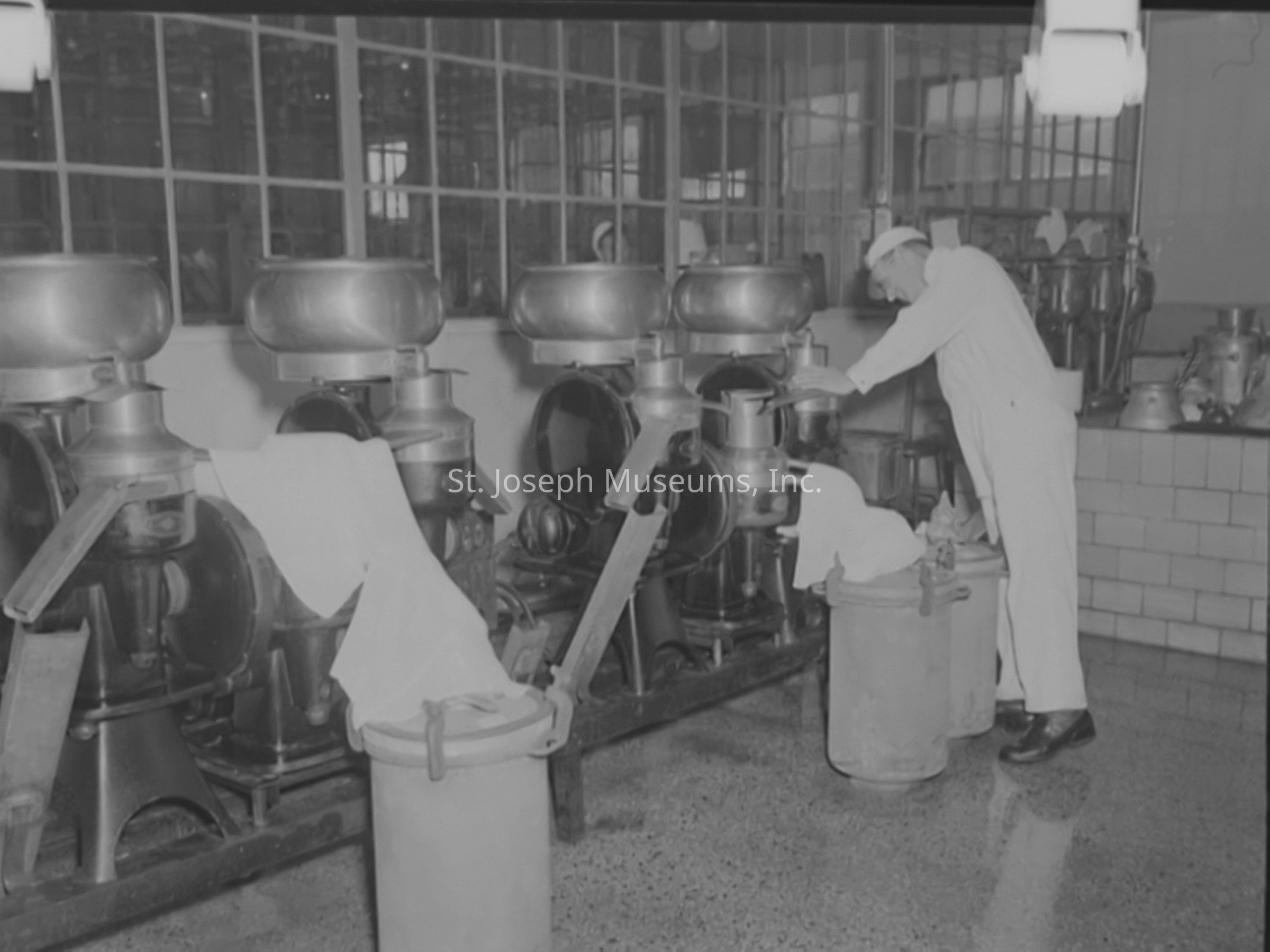 Black-and-white image showcasing a worker dressed in white interacting with machinery inside the Anchor Serum Company. Industrial equipment, such as large mixers or processors, lines the room. The floor is polished and clean, reflecting the machinery above. In the background, windows reveal additional spaces suggesting a larger industrial facility.