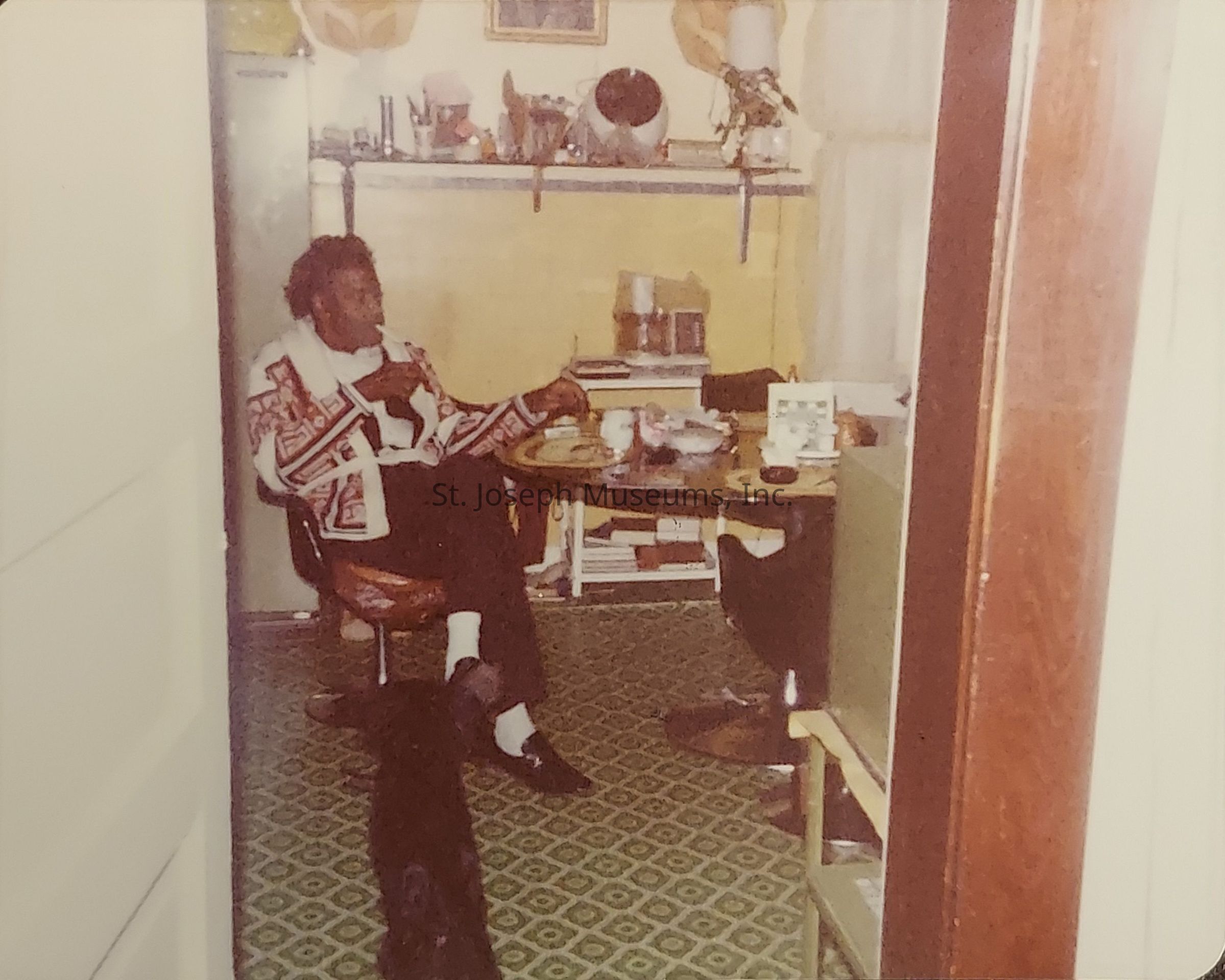 The image depicts a person seated at a round table covered with various items, in a room with patterned green carpet flooring. They are wearing a patterned sweater and appear to be engaged in eating or drinking, holding a cup or glass in one hand while looking to the side. The room's walls are adorned with shelves containing different objects, including a framed picture and a lamp that hangs above. A fridge is visible in the background near the left corner. The person's foot rests on the floor, and there appears to be a black dog lying down on the carpet nearby. The scene seems intimate and relaxed, as if capturing a candid moment at home, viewed through an open door leading into the room, bordered by wooden frames, adding a cozy atmosphere.