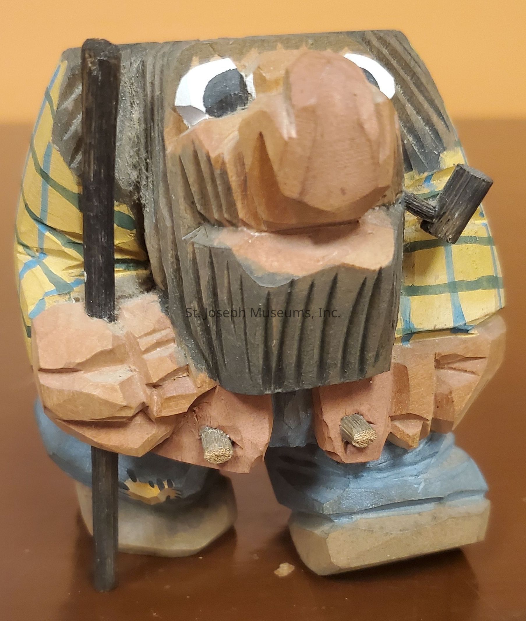 The image displays a small, handcrafted wooden figurine characterized by exaggerated features. The figure has a large nose and wide eyes, giving it a whimsical appearance. It features a thick, textured beard and is dressed in a yellow plaid shirt, suggesting a rustic or outdoorsy theme. The shirt contrasts with its blue jeans and tan shoes. The figure is bent slightly, holding a walking stick in its left hand, adding a sense of movement or purpose. The wood carving shows visible chisel marks, indicating a detailed, artisanal creation. Its overall design combines elements of humor and craftsmanship, capturing a charming, rustic character.