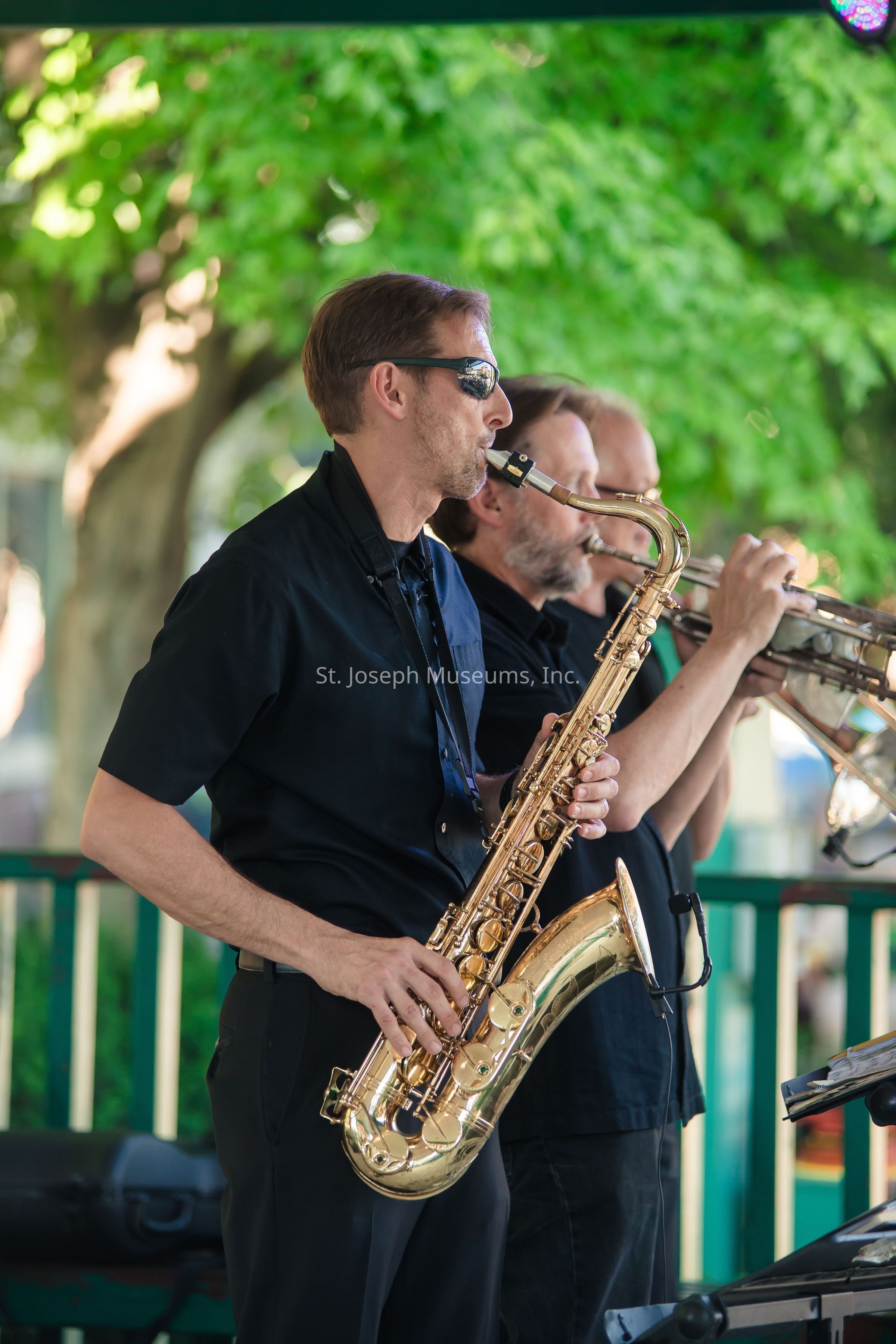 In the image, a group of musicians performs outdoors, with a focus on a saxophonist playing his instrument passionately while wearing sunglasses and dressed in a black shirt, standing alongside another musician playing a trumpet; the background showcases vibrant green trees indicating a lush environment, which contrasts with the musicians' dark attire, creating a vivid scene of a live musical performance that evokes a sense of rhythm and melody, as the players appear to be deeply engrossed in their music, with the sunlight filtering through the leaves adding a natural spotlight effect that highlights the energy and ambiance of the setting.
