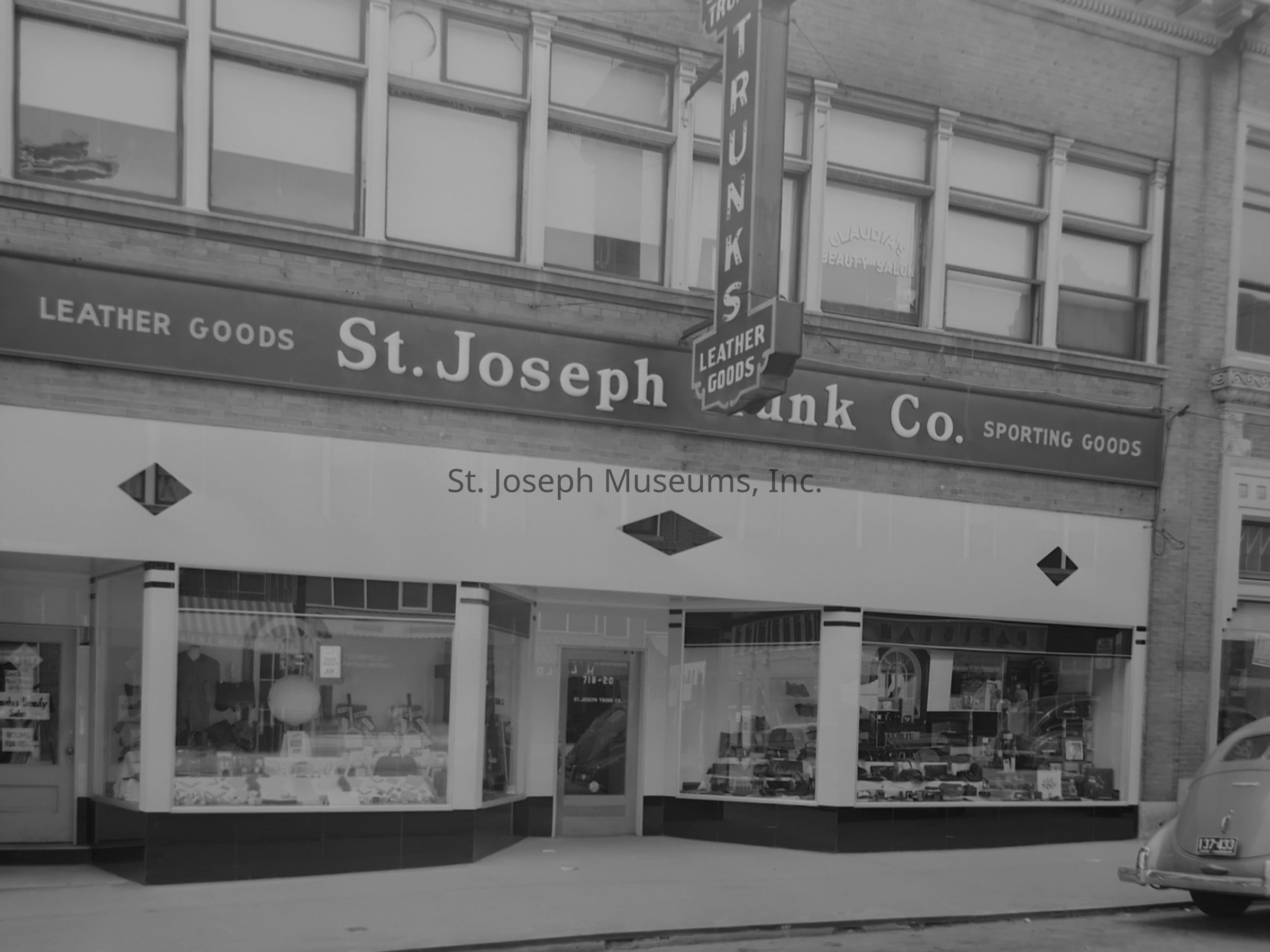 Black-and-white image of the exterior of St. Joseph Trunk Co., located at 418-420 Francis St., showcasing a storefront with large display windows filled with various leather and sporting goods. Above the windows, a prominent sign reads "St. Joseph Trunk Co." accompanied by "LEATHER GOODS" and "SPORTING GOODS." A vertical sign protrudes from the building, labeled "TRUNKS" and "LEATHER GOODS."