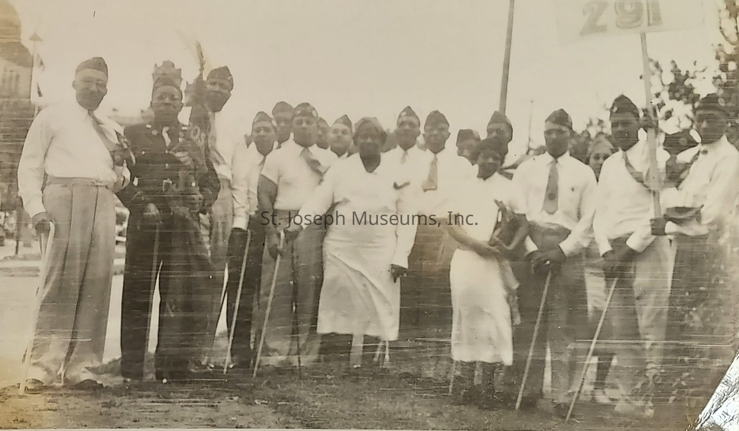 The image presents a group of people, likely veterans or members of a formal organization, gathered for a photo. They are wearing uniforms or formal attire, consisting of dark jackets, light trousers, and ties, with most holding canes or walking sticks. A mix of men and women stand closely together, and some wear hats. In the background, there's a flag with the number "291," along with some trees and a building with a dome-shaped roof, adding context to the setting. The image has an aged, sepia tone, suggesting it is from a past era, possibly mid-20th century, evoking a sense of history and camaraderie.