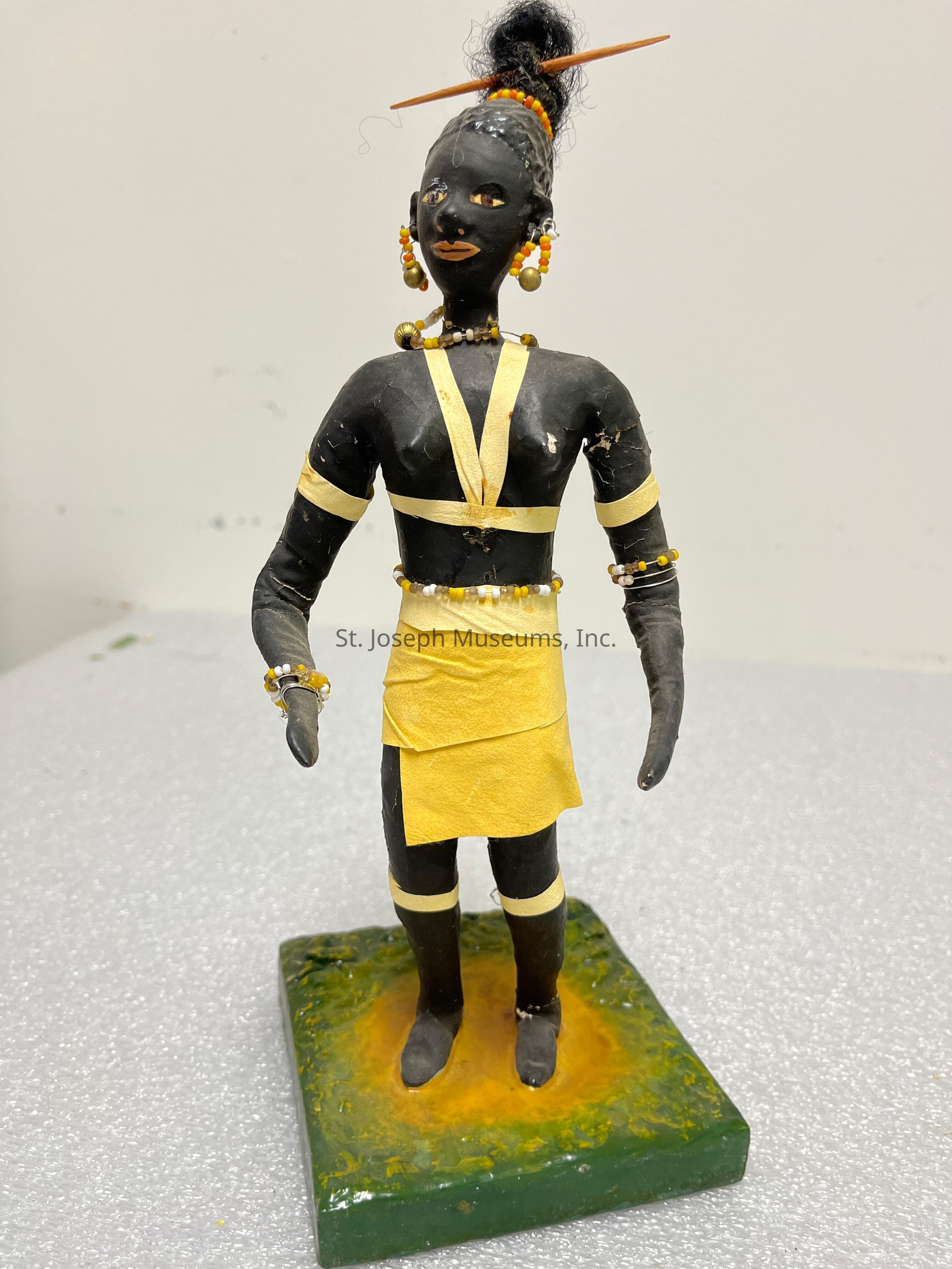 The image showcases a handcrafted figurine, representing a person with dark skin and adorned in traditional attire. The figure is clothed in a yellow skirt and matching bands that wrap around its arms and legs. It is accessorized with beaded jewelry, including orange and yellow beads that form earrings, a necklace, and bracelets. The hair is styled in an updo, secured with a stick adorned with beads, and the whole figure is positioned on a square, two-toned base with green and yellow hues. The craftsmanship and attention to detail highlight cultural and artistic expression.