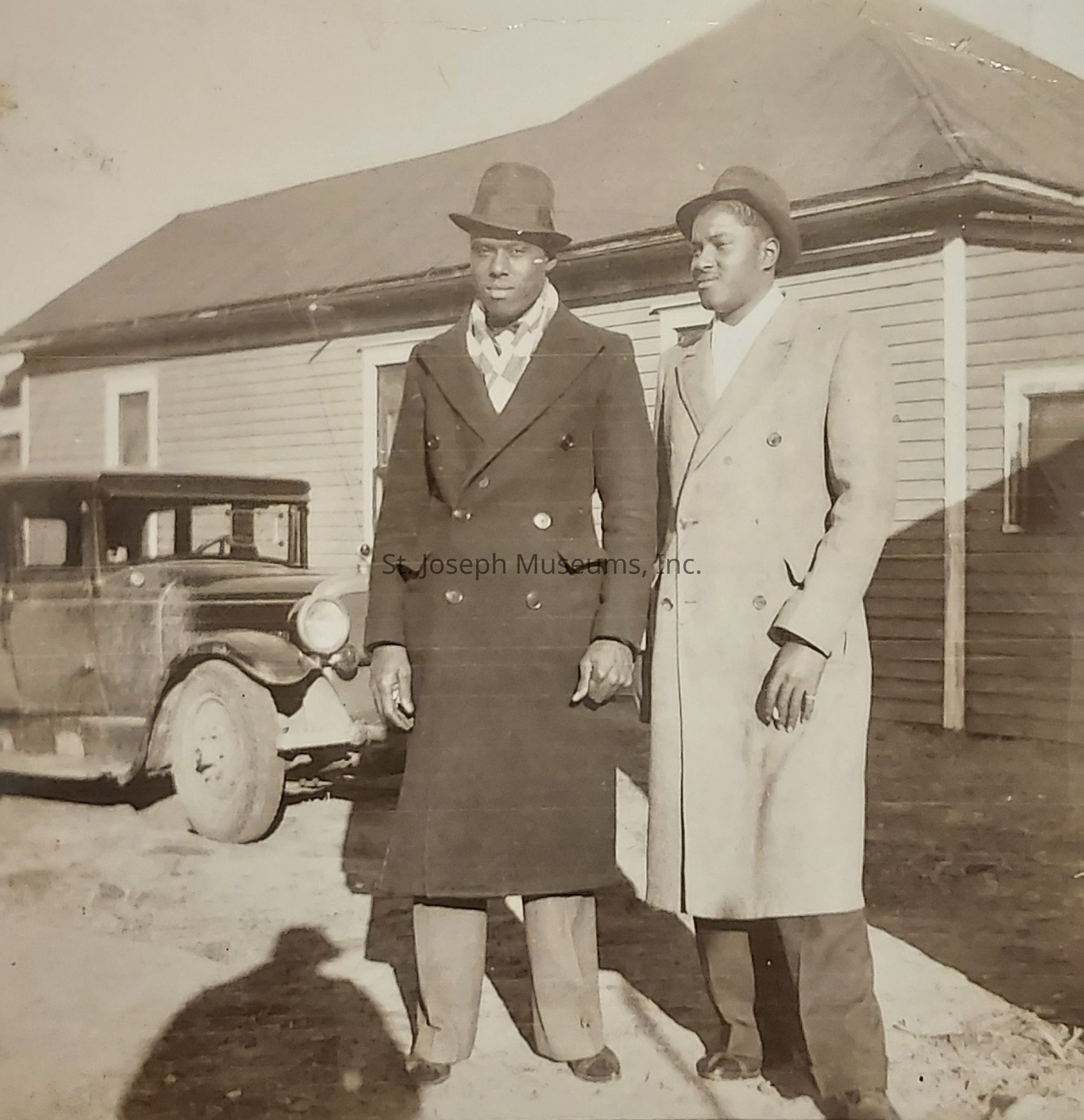 The image depicts two men dressed in well-tailored, double-breasted coats, each wearing a hat, standing side by side in front of a vintage car. They are positioned in front of a wooden house, suggesting a setting from a bygone era. The scene is in black and white, enhancing the historical feel. Shadows on the ground indicate it is a sunny day. The men appear composed and dignified, exuding an air of confidence from their stance and attire. The car behind them has classic features, with spoked wheels and visible headlamps, further emphasizing the image’s period context. The house has a simple, rustic design, with horizontal planks and a sharply pitched roof, typical of early 20th-century architecture. The men's attire, including their hats and overcoats, reflects a formal style, possibly indicating the photograph was taken for a significant occasion or event. The overall atmosphere captured in the image evokes a sense of nostalgia, offering a glimpse into the past.
