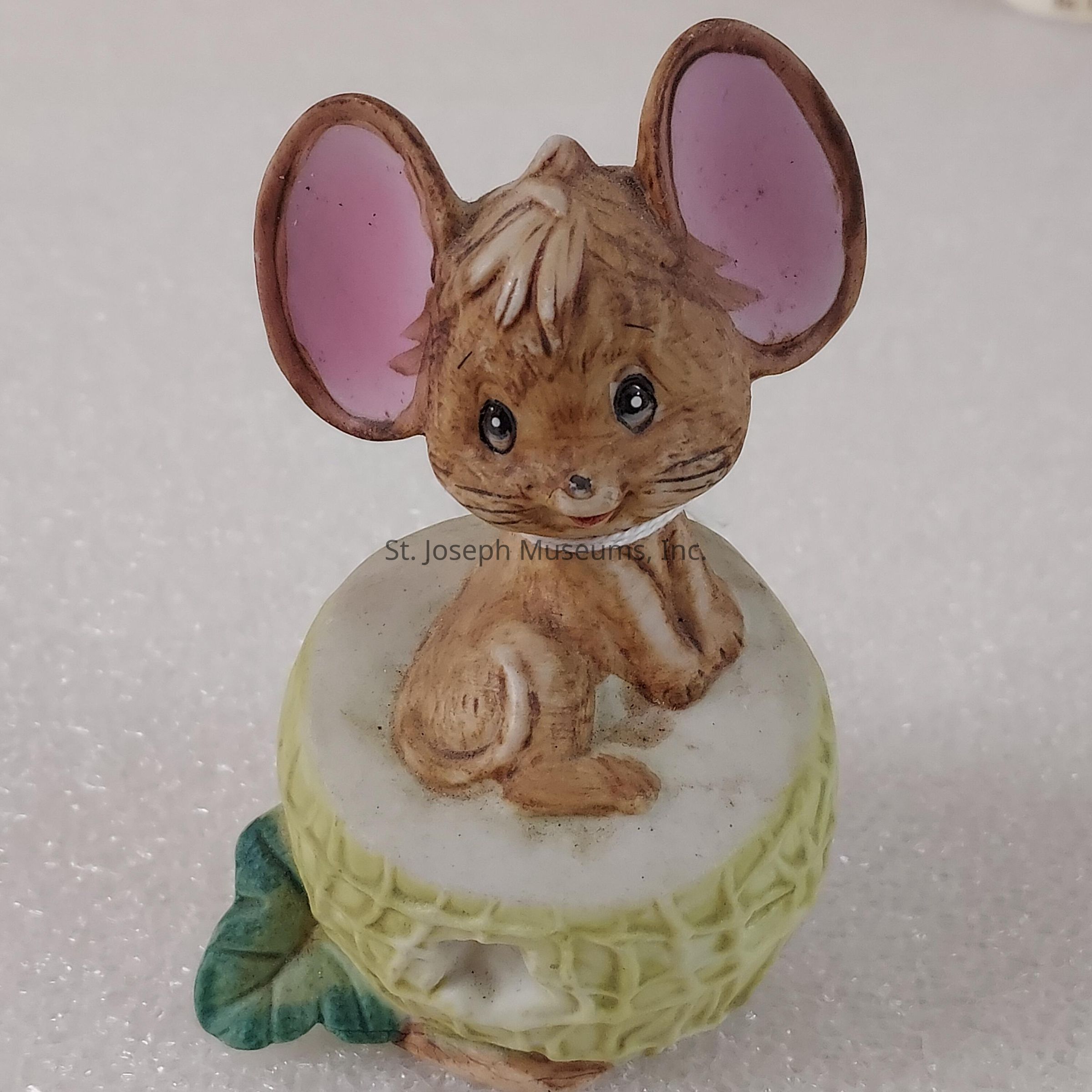 The image depicts a charming figurine of a cute, anthropomorphic mouse with large, expressive ears and detailed fur texture, sitting on what appears to be a small, round object resembling a piece of pale yellow cheese or a similar textured item. The mouse is depicted in a sitting posture, seemingly at ease, and features an endearing facial expression with wide, sparkling eyes, adding to its adorable appearance. Its inner ears are painted a soft pink, contrasting with the brown fur, which is carefully shaded to highlight the texture. Behind the mouse, there's a green leaf accent at the base, likely adding an element of nature to the composition. The background consists of a soft, neutral surface, allowing the figurine to stand out prominently, emphasizing its intricate design and the artist's attention to detail. The piece is crafted with a subtle color palette that enhances the figurine's whimsical and friendly aura.