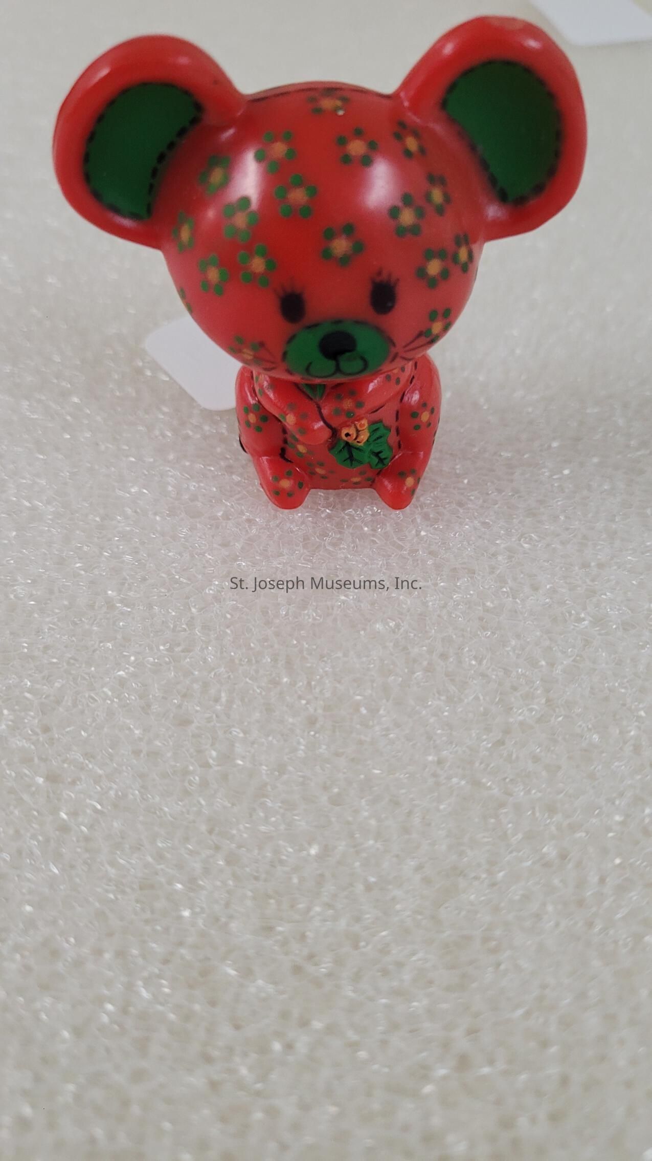 The image showcases a small, decorative figure resembling a mouse, characterized by its vibrant red color adorned with small green floral patterns across its entire body. The green accents are prominently visible in the inner parts of its large, round ears and its snout, adding contrast to the predominantly red surface. Its eyes and nose are simplistic yet cute, crafted with basic black lines that give it a friendly and playful expression. This figure is resting on a textured, white bubble wrap surface, which provides a neutral background that accentuates the bright colors of the figure itself. The collectible item is likely made from a glossy material, such as plastic or resin, as indicated by the sheen that reflects light and highlights the contours of the figure. This whimsical ornament could serve as a charming piece for collectors or anyone with an affinity for cute and quirky designs.